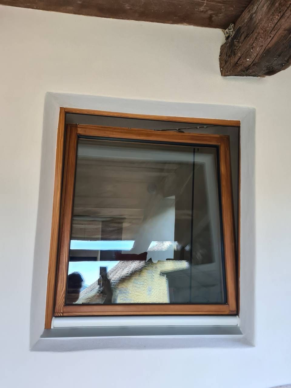 Finestra quadrata, aperta, con cornice in legno, su una parete bianca; vista esterna riflessa nel vetro.