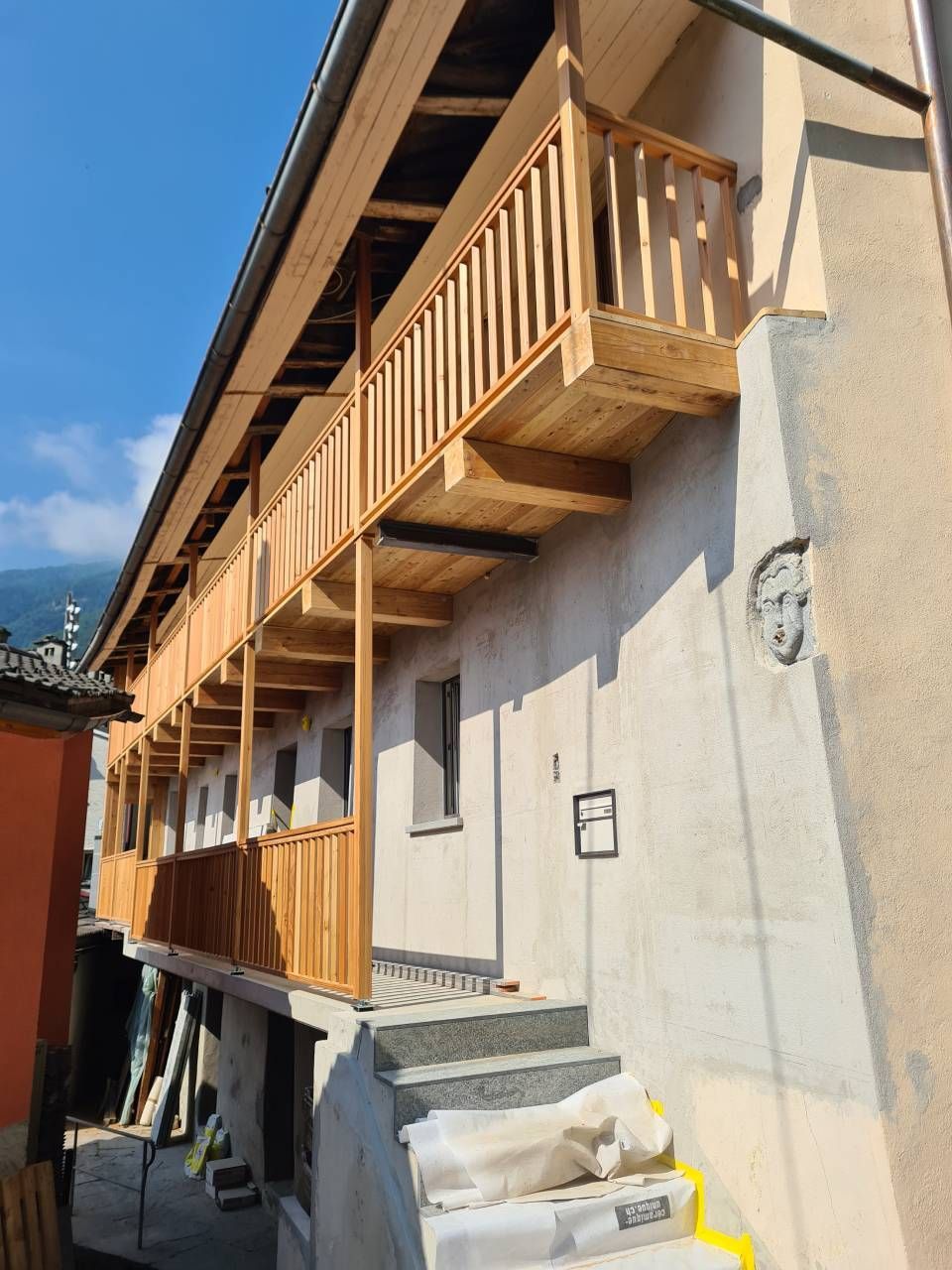 Balcone in legno su un edificio chiaro con scale che portano al piano superiore e cielo azzurro sullo sfondo.
