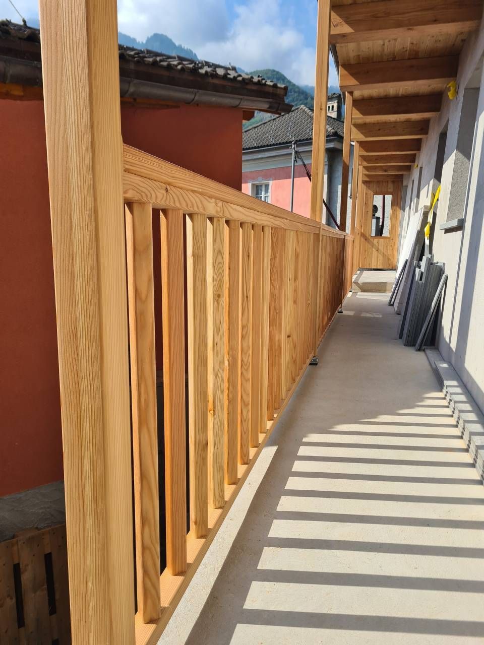 Ringhiera in legno su uno stretto passaggio esterno. Soleggiato con vista su edifici e montagne.