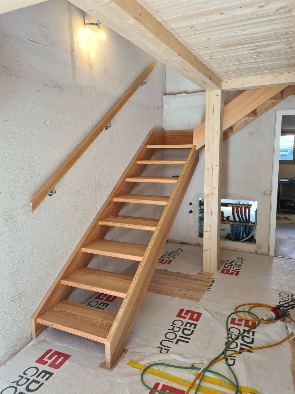 Scala in legno con corrimano in una casa che conduce a una trave e a una stanza con luce naturale.