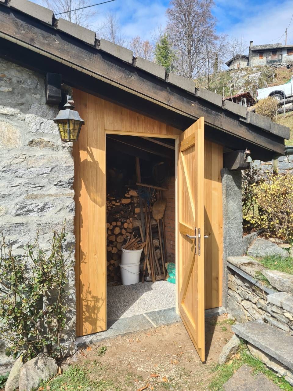 Capanno in legno con porta aperta che rivela legna da ardere e pale. Esterno in pietra con tetto sporgente.