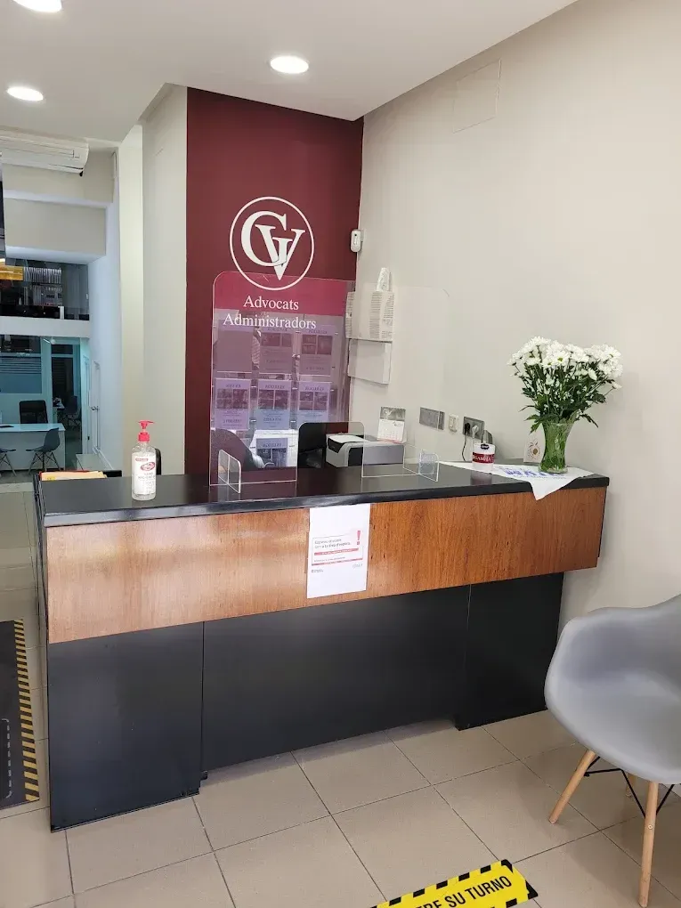 Mostrador de recepción con logotipo en la pared. Se ven una botella de desinfectante de manos y un ramo de flores blancas.