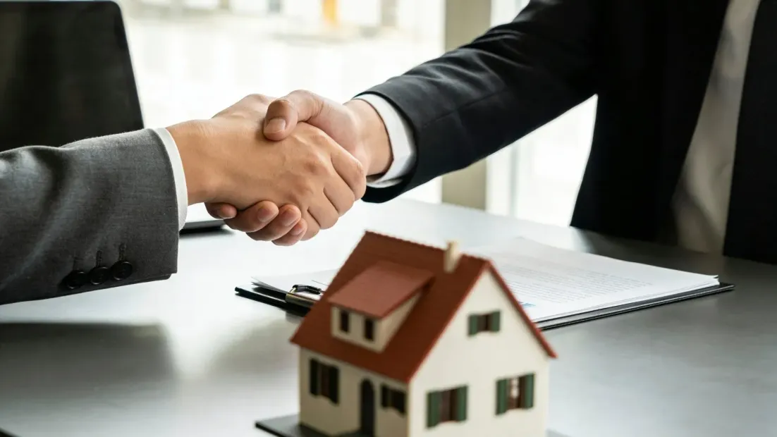 Dos personas se dan la mano sobre una casa en miniatura, simbolizando un acuerdo inmobiliario.