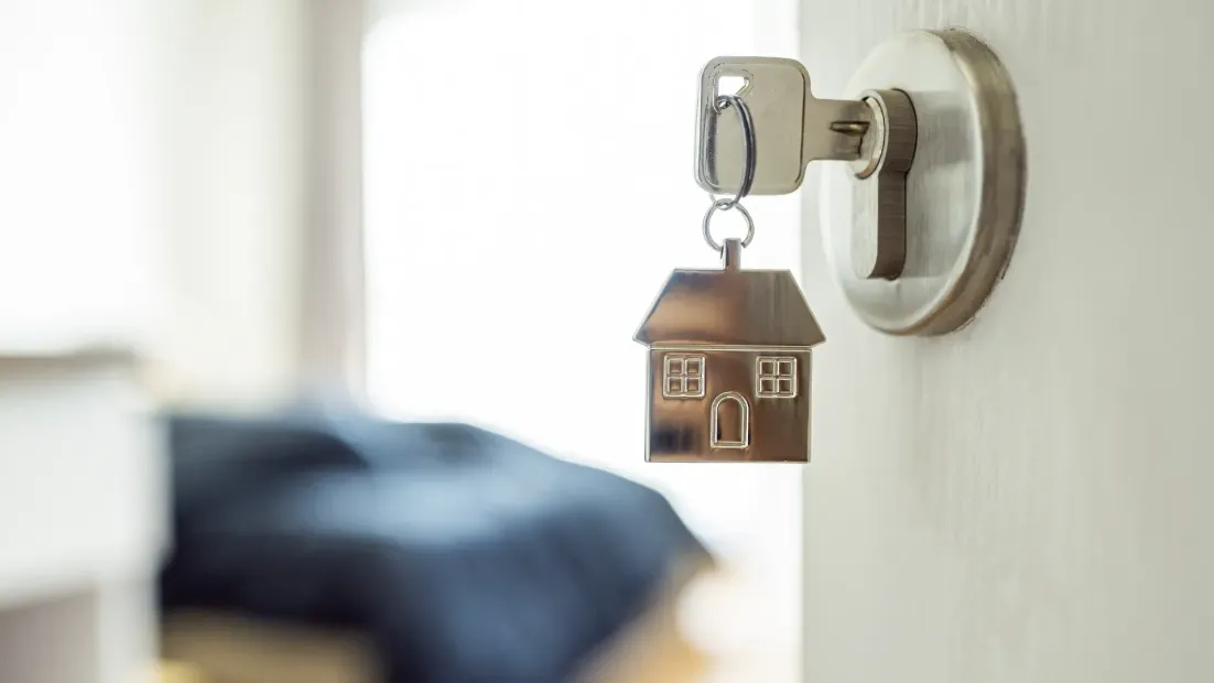 Entra en una puerta con un llavero con forma de casa, con un dormitorio borroso visible al fondo.