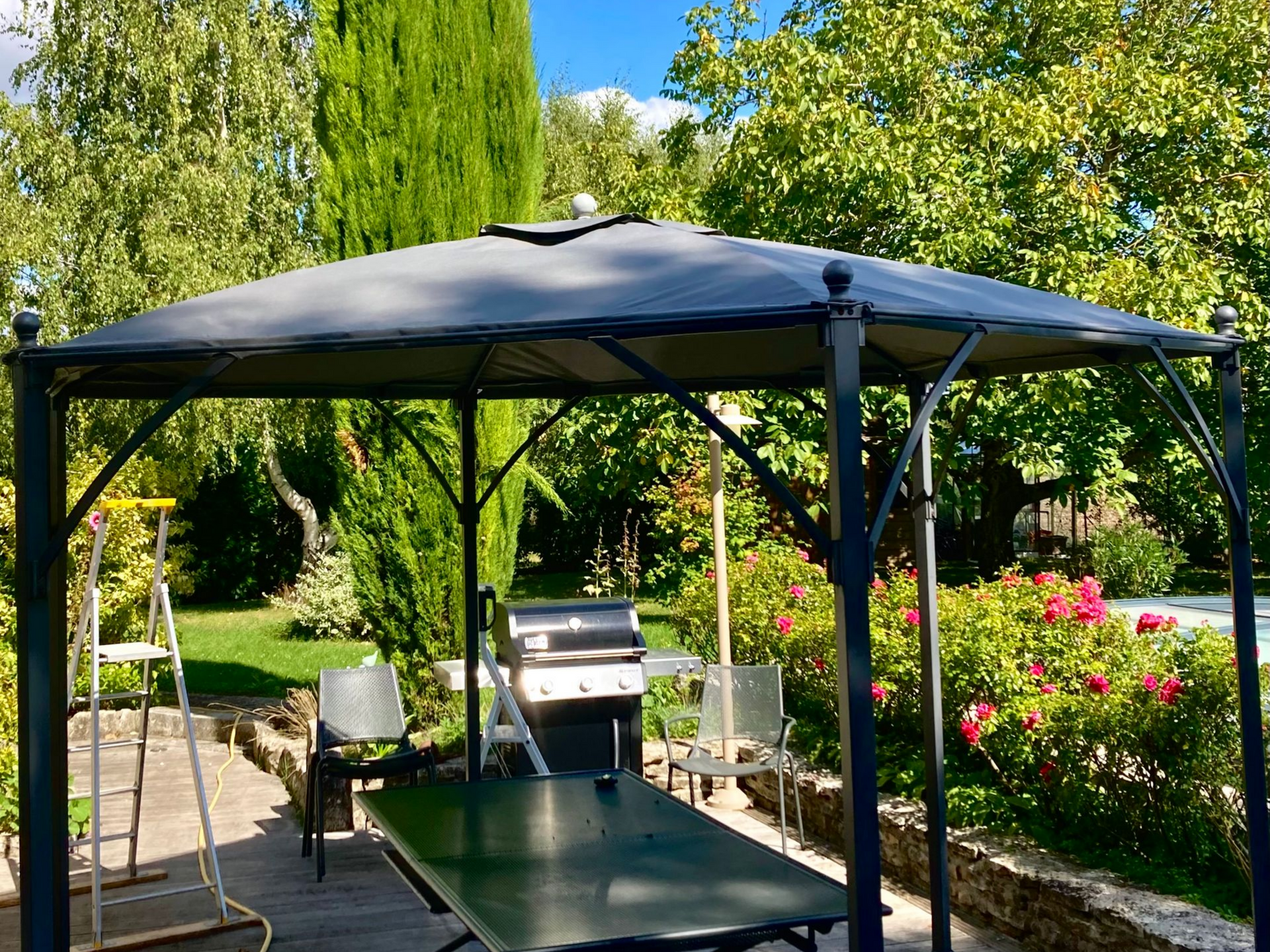 Une gloriette à la structure sombre se dresse sur une terrasse avec une table, entourée d'un jardin verdoyant luxuriant.