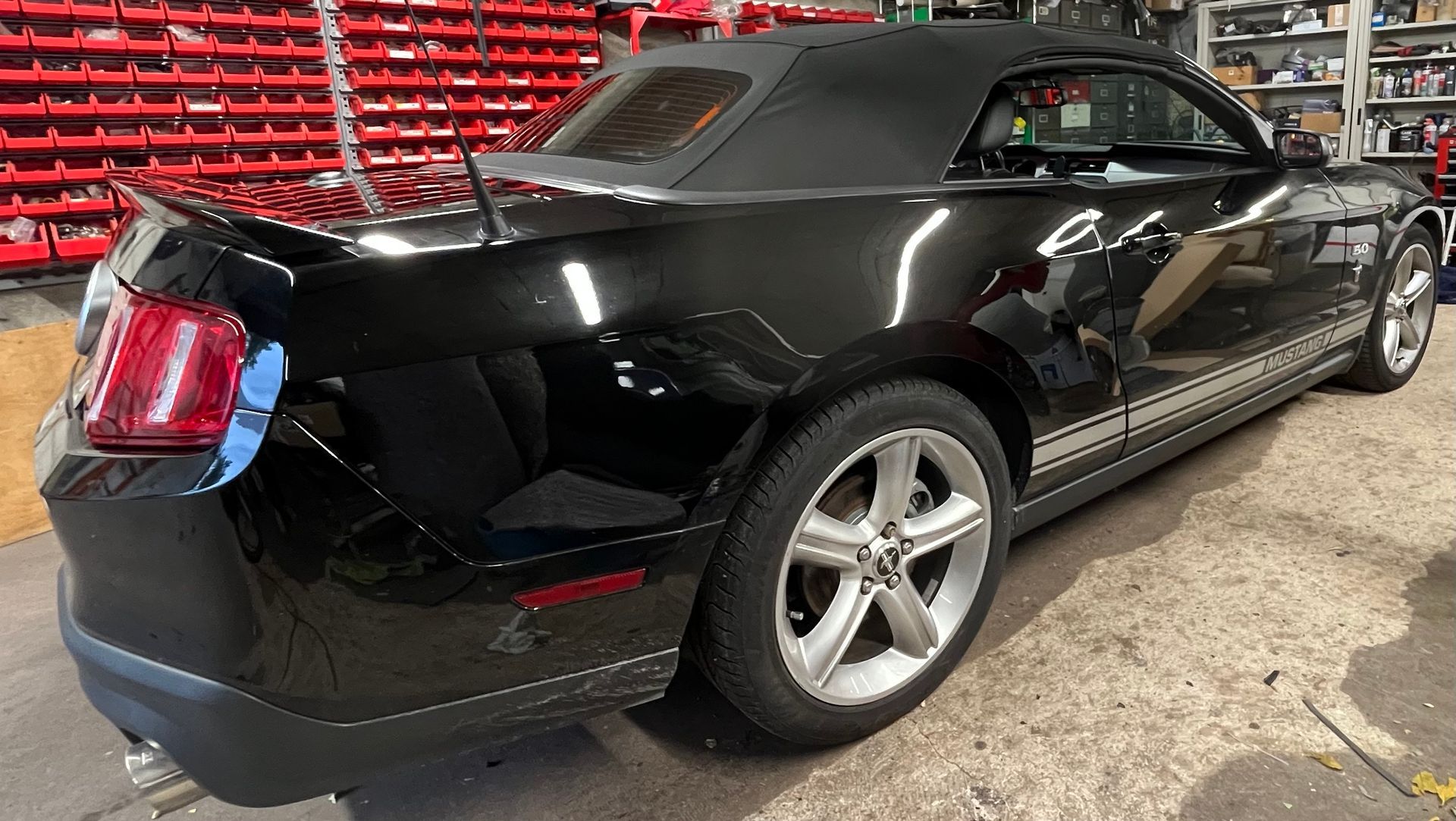 Une Ford Mustang décapotable noire garée dans un garage, avec des bacs de rangement rouges en arrière-plan.