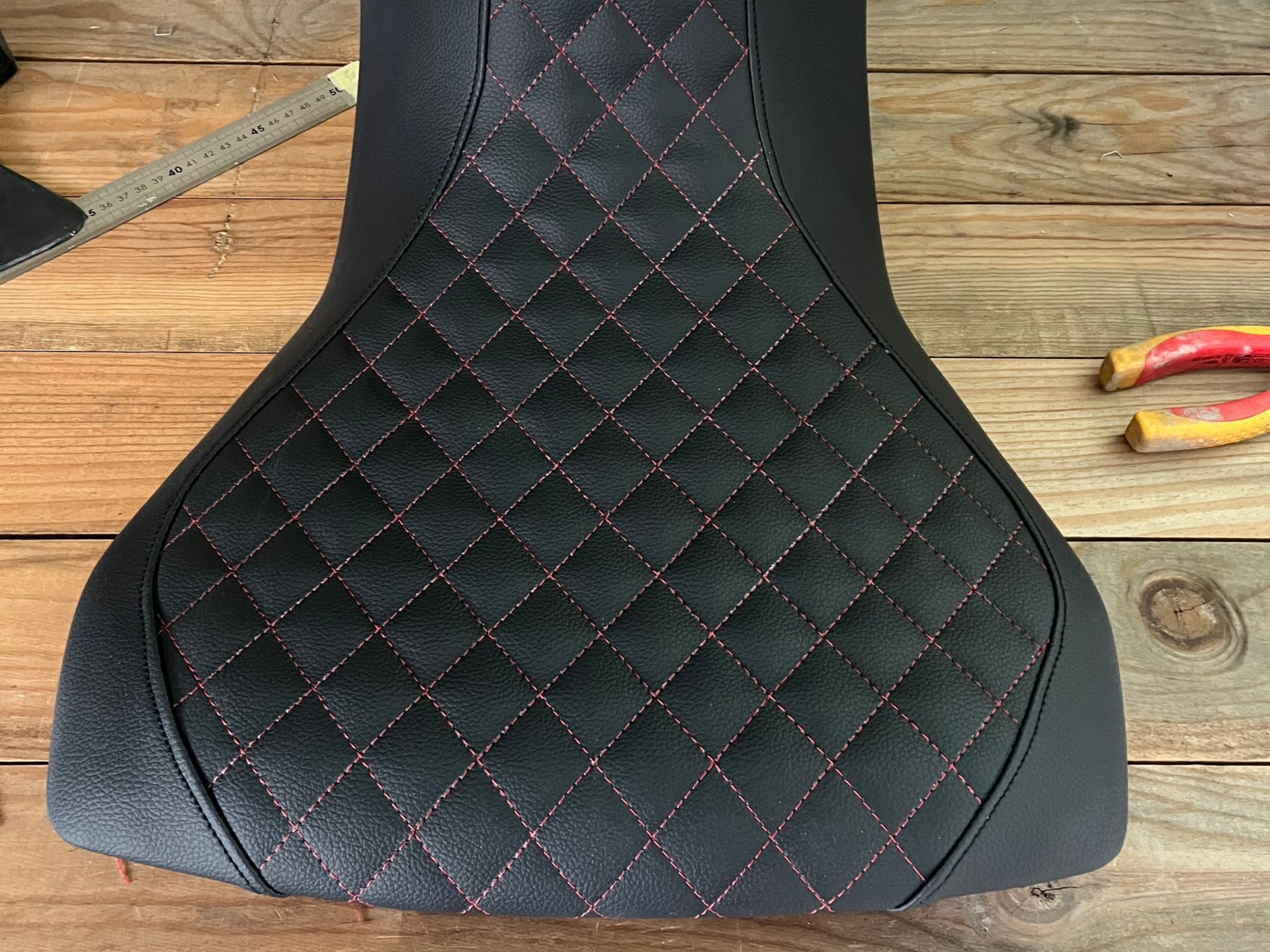 Selle de moto noire à motifs de losange rouges, posée sur une surface en bois rustique à côté d'outils.