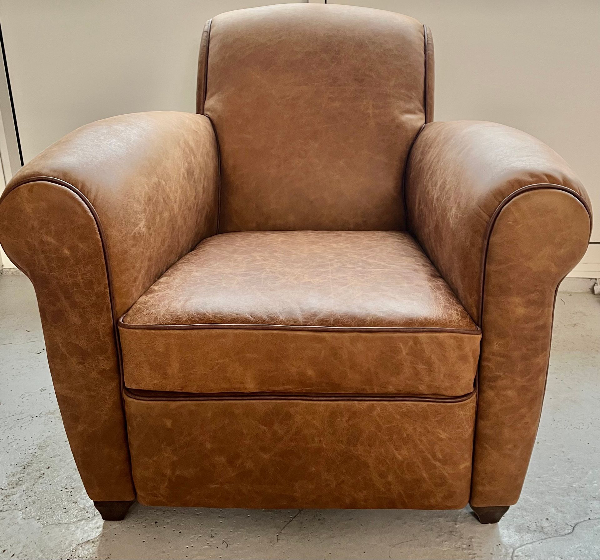 Un fauteuil club en cuir beige aux accoudoirs arrondis, posé sur un sol blanc contre un mur uni.
