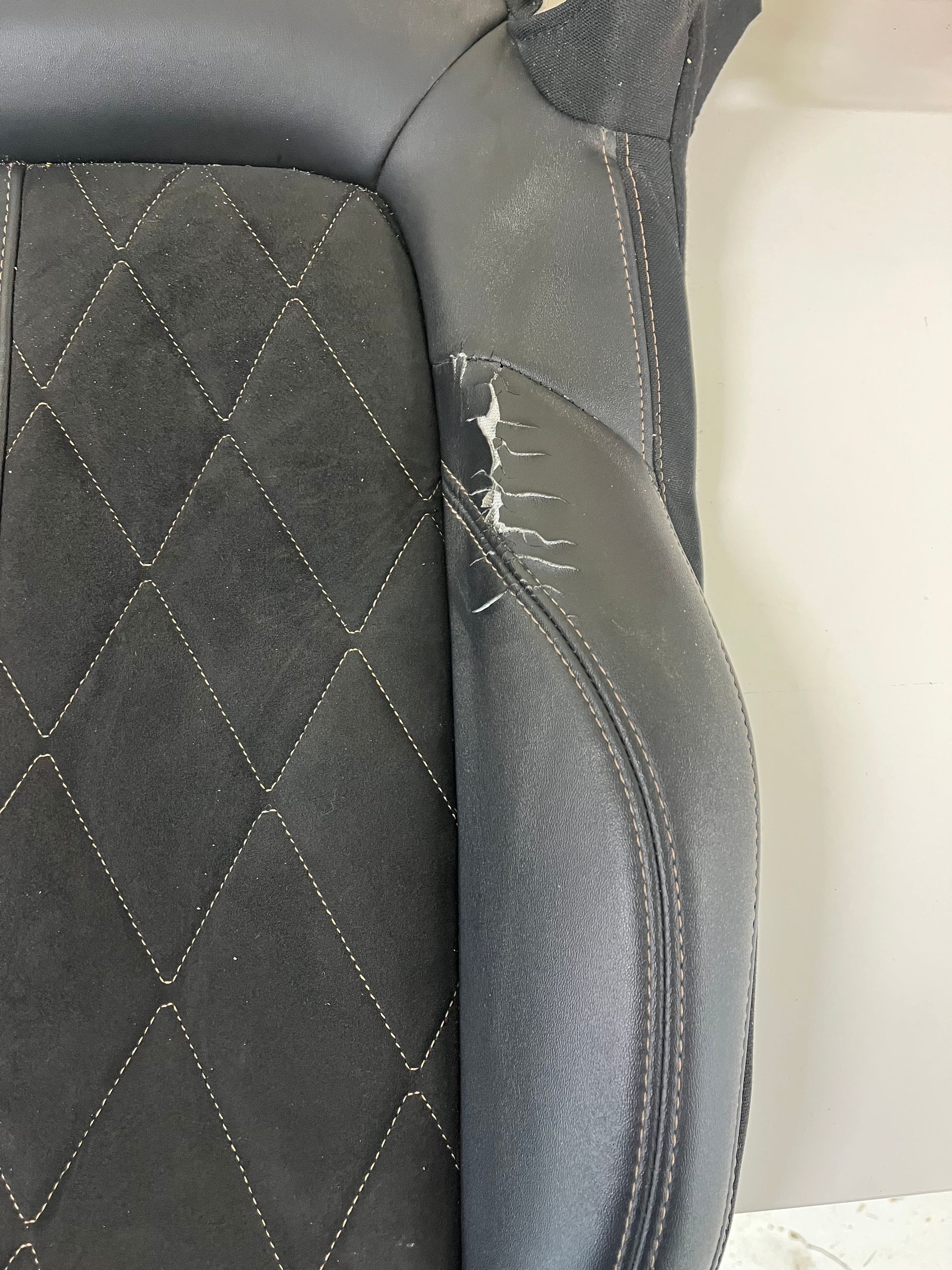 Un siège auto noir avec un centre en daim surpiqué en losanges et un renfort en cuir abîmé dont la surface se décolle.