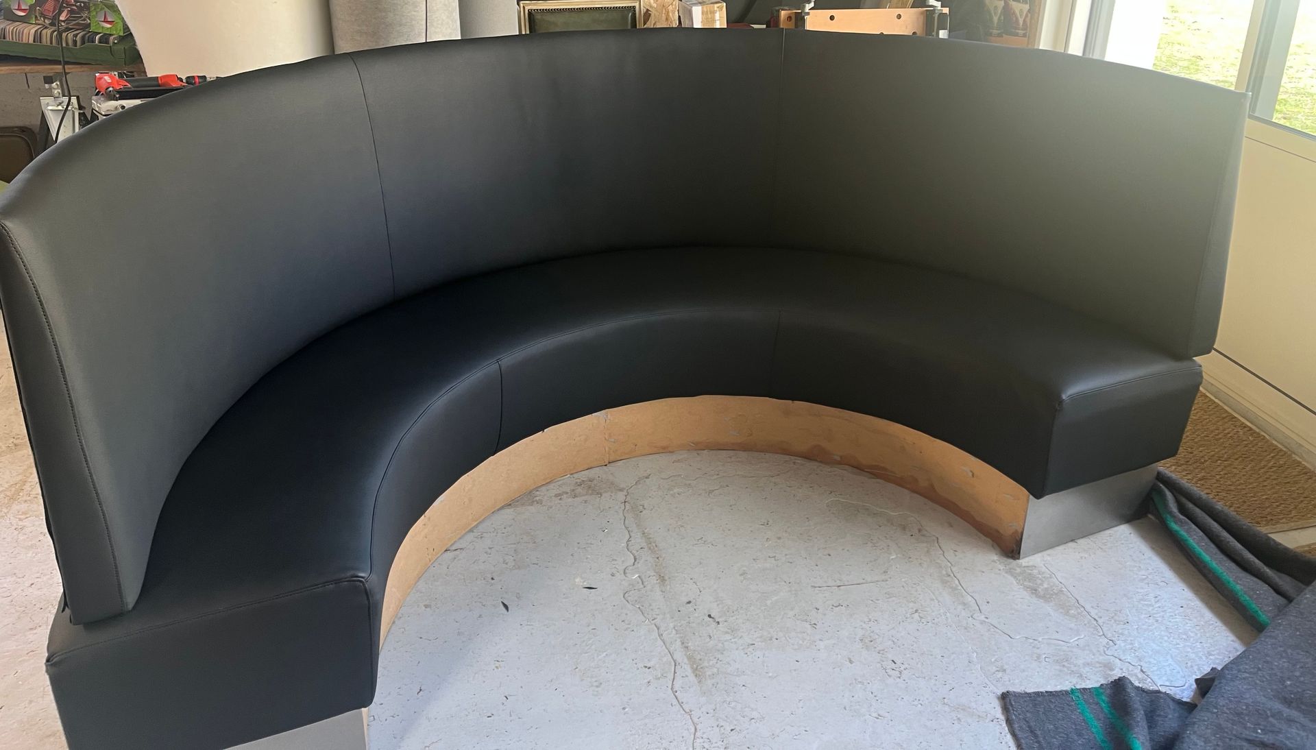 Dans une pièce inachevée, une banquette incurvée en cuir noir, avec un socle en bois et des pieds argentés, est installée.