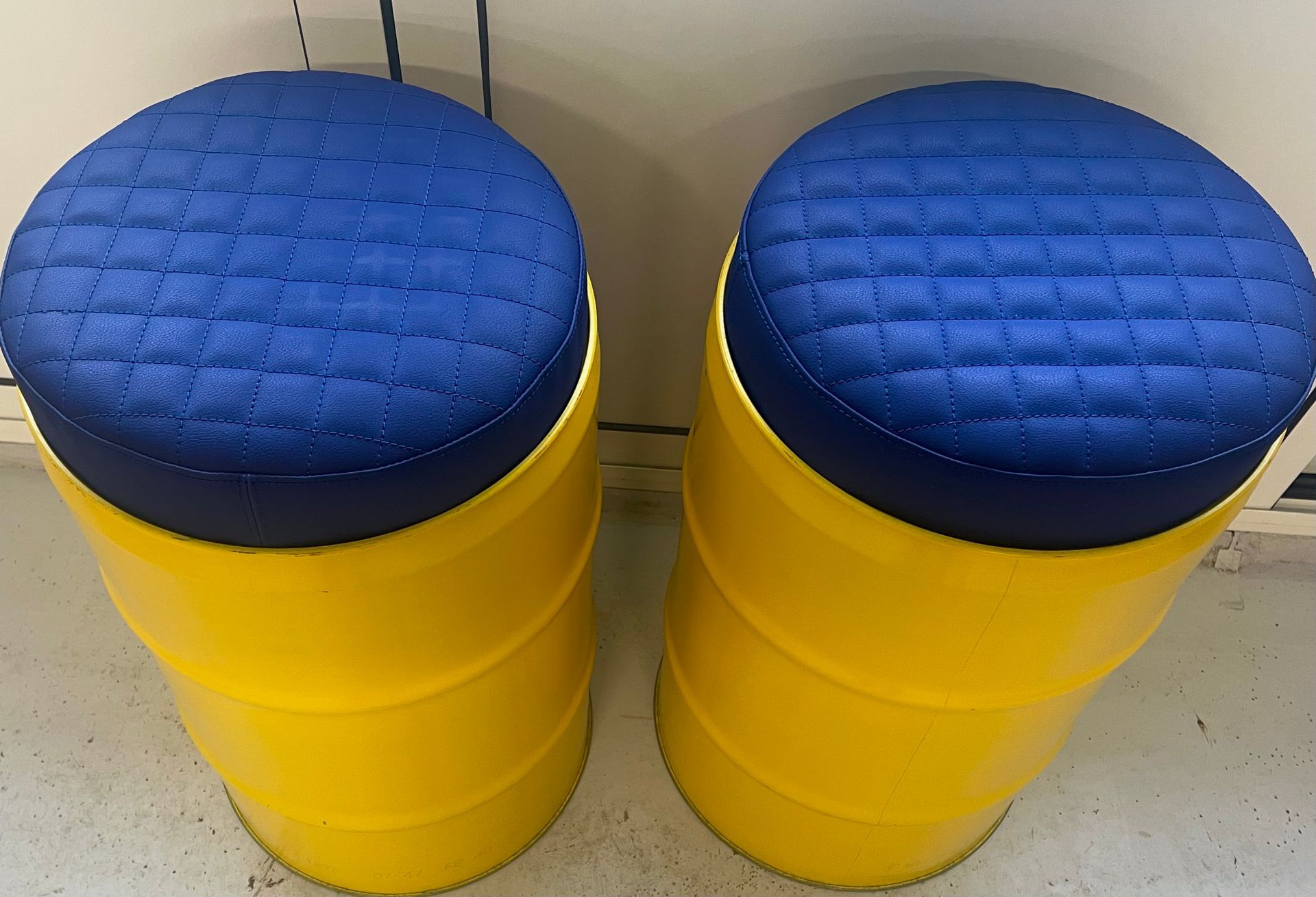Deux tabourets fabriqués à partir de fûts en métal jaune, avec des assises rondes, matelassées et rembourrées de bleu.