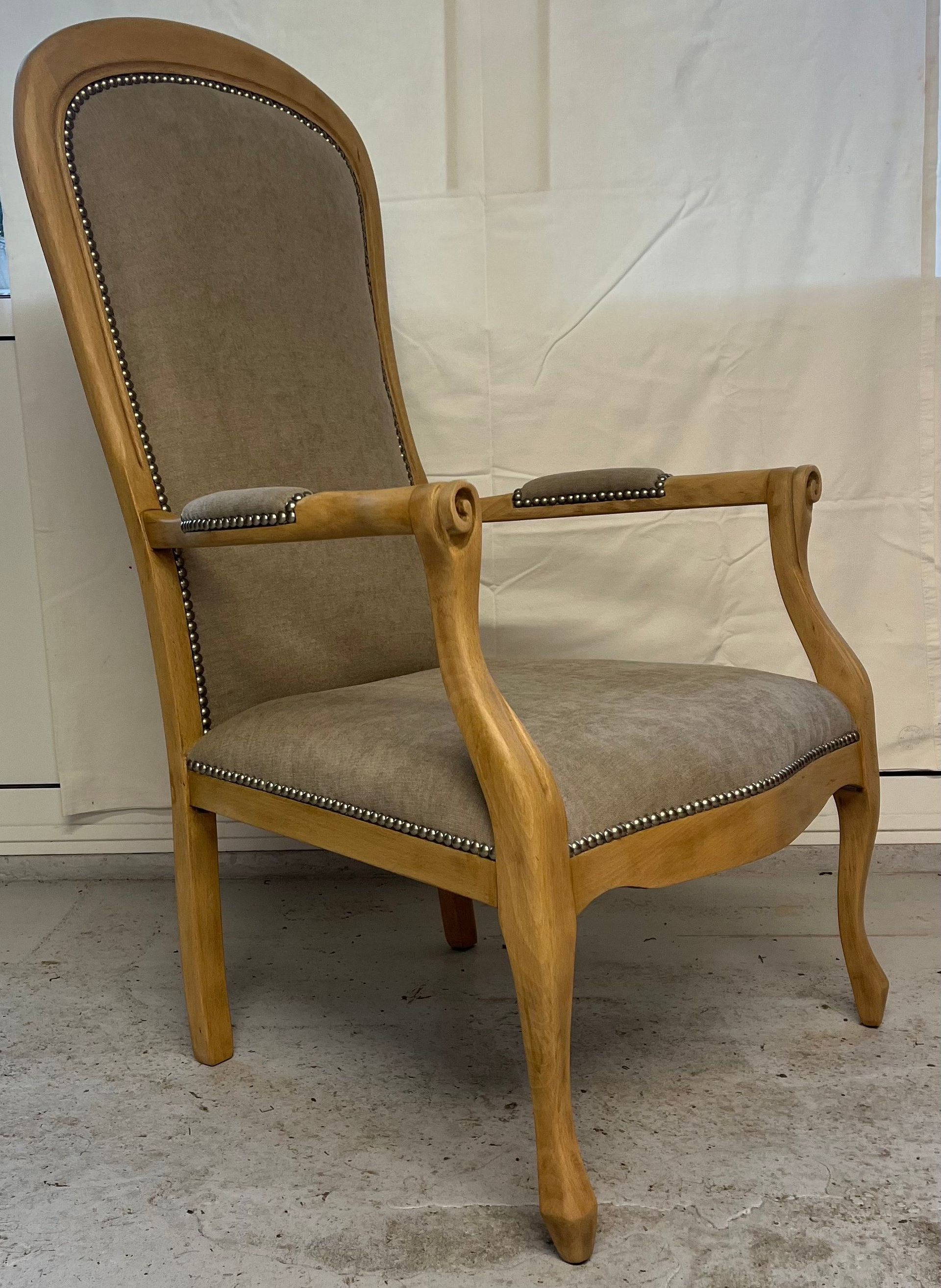 Un fauteuil haut en bois clair, avec assise et dossier rembourrés gris neutre, orné de clous décoratifs.