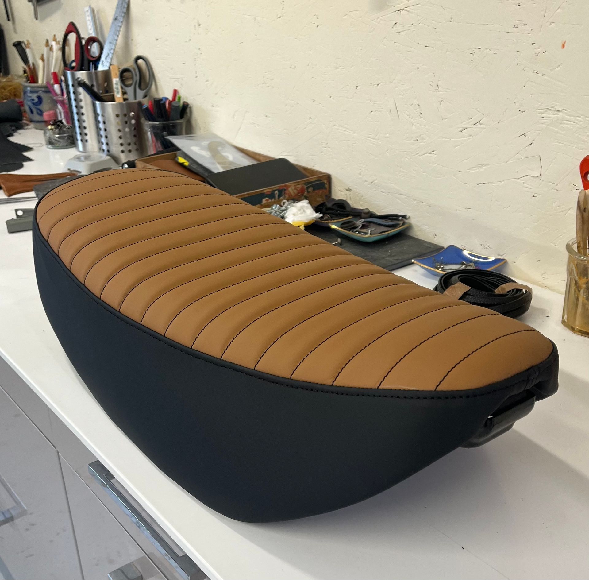 Une selle de moto personnalisée, avec un dessus côtelé beige et des côtés noirs, posée sur un établi dans un atelier.