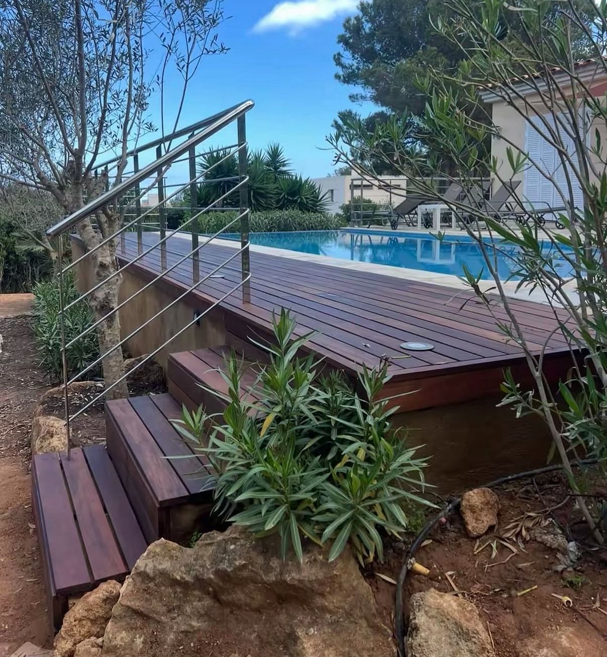 Terraza y escalones de madera que conducen a una piscina enterrada, barandilla de acero inoxidable, entorno exterior soleado.