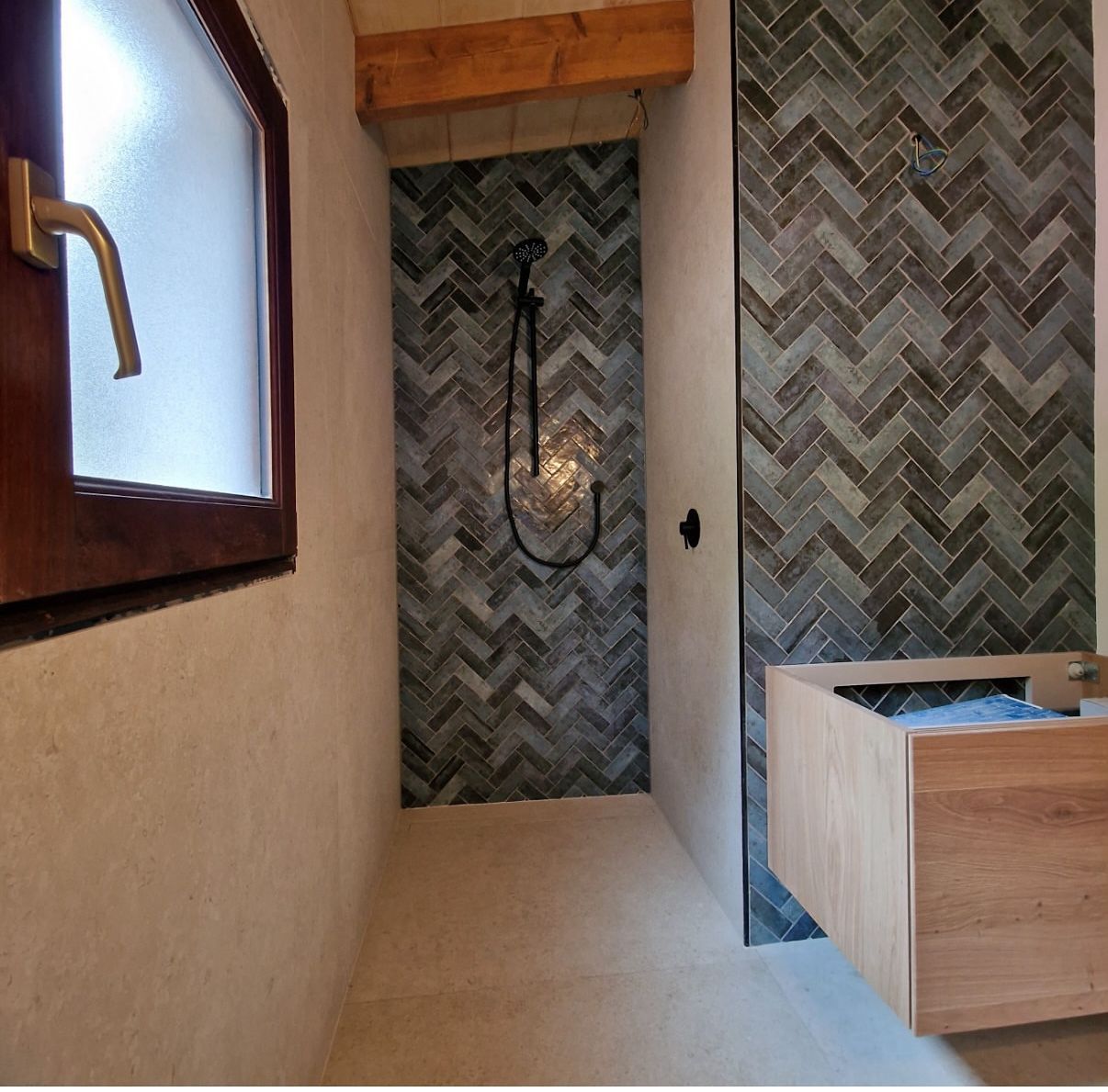 Baño pequeño con ducha de azulejos en espiga, tocador de madera y ventana con herrajes dorados.