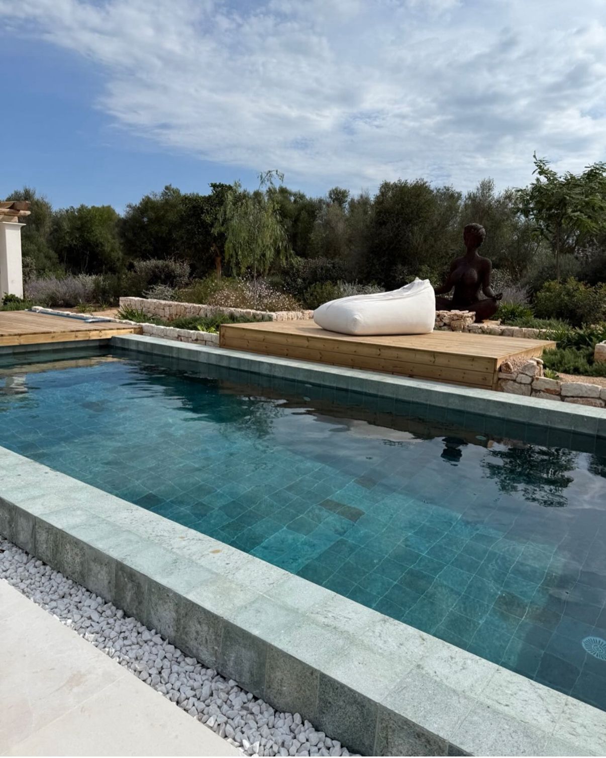 Piscina de agua azul verdosa, puf blanco sobre plataforma de madera, escultura en un entorno natural al aire libre bajo un cielo parcialmente nublado.