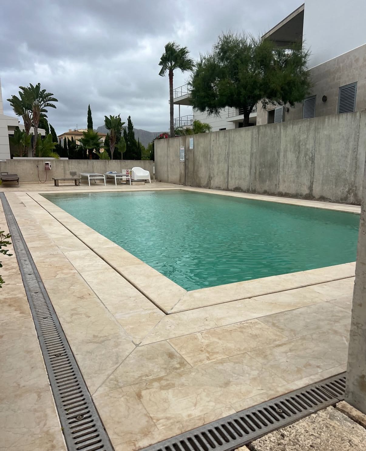 Piscina rodeada de piedra clara. Rejillas de drenaje grises y muros de hormigón. Cielo nublado.