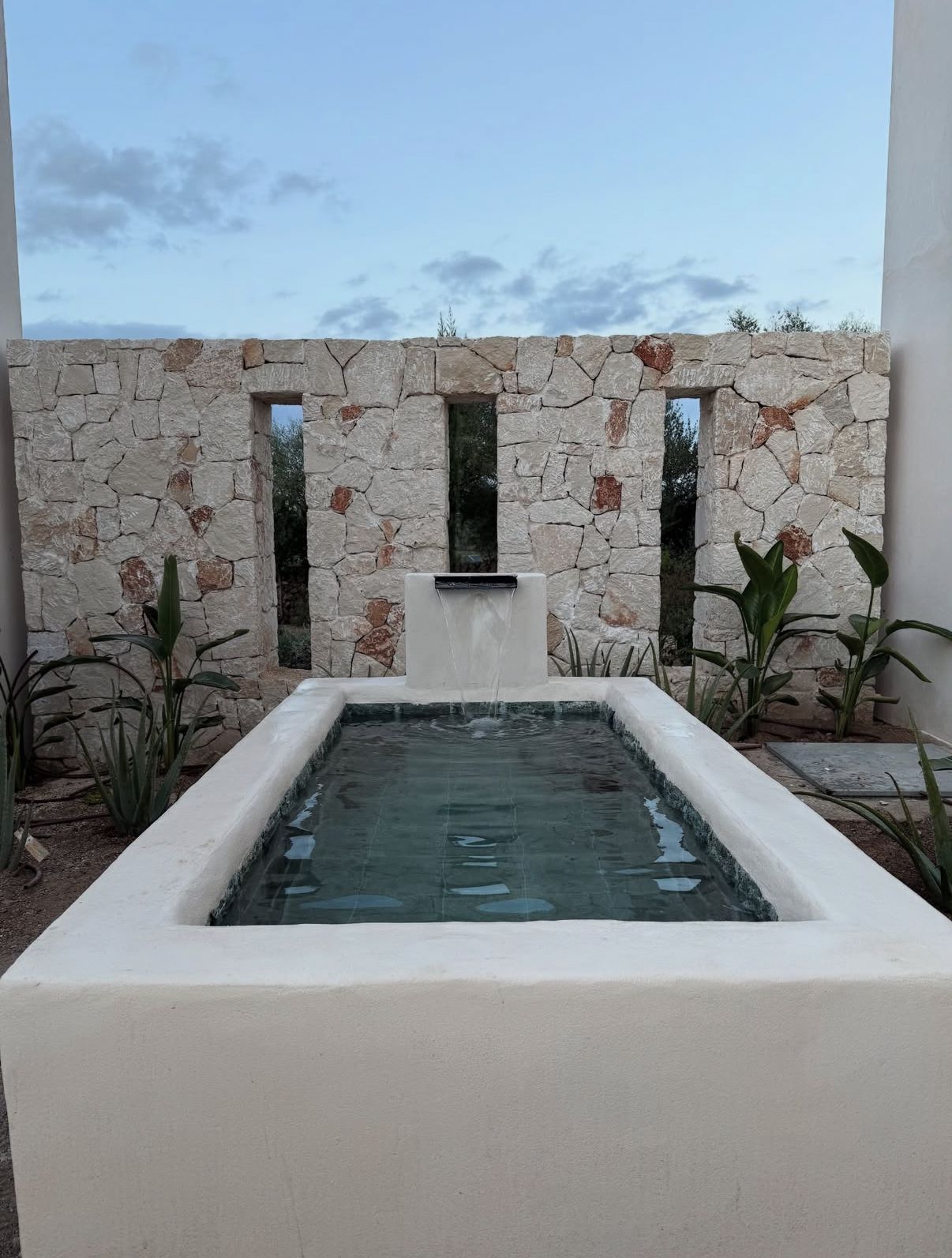 Muro de piedra con tres aberturas sobre una piscina blanca, rodeada de plantas. Cielo al fondo.