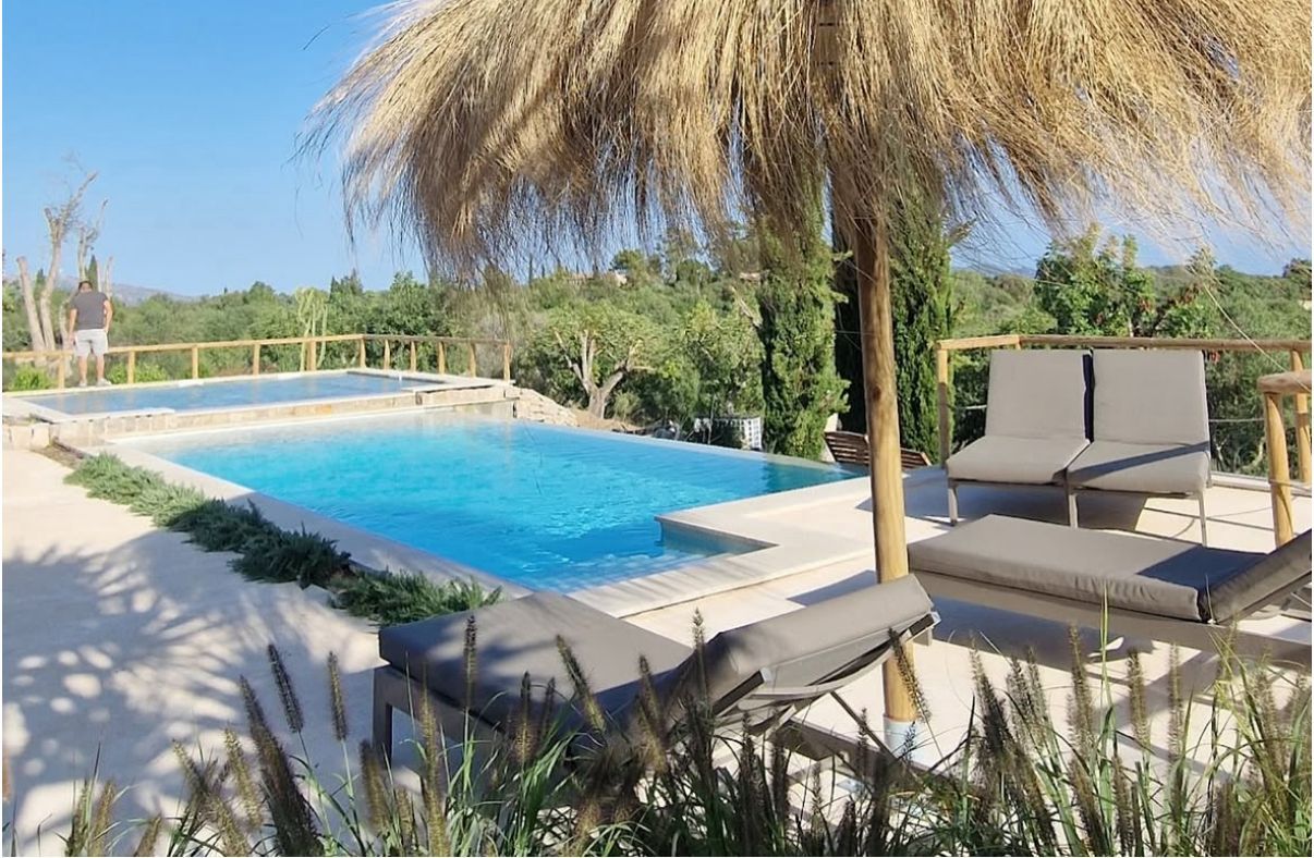 Piscina con tumbonas bajo una sombrilla de paja, con vistas a un paisaje verde.