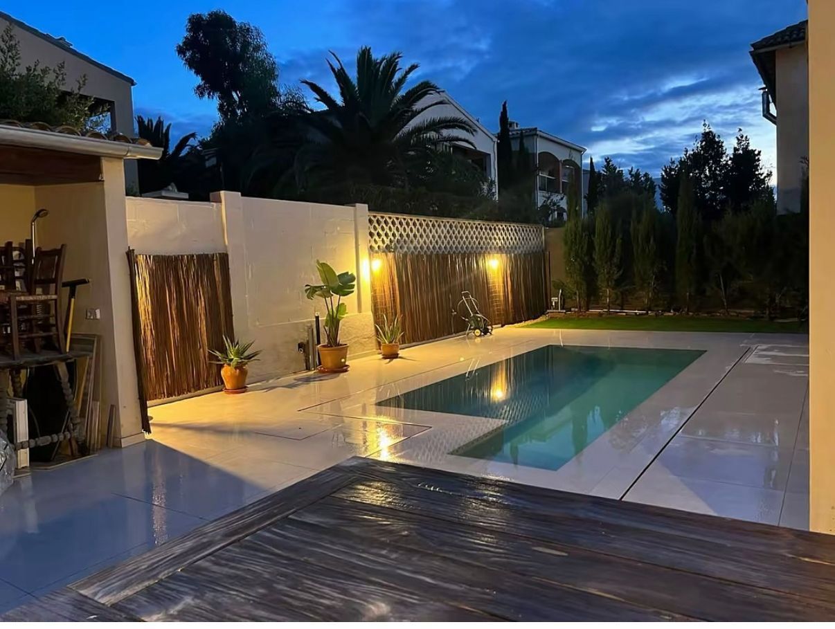 Vista nocturna de una zona de piscina con paredes iluminadas, una palmera y una piscina con agua reflectante.