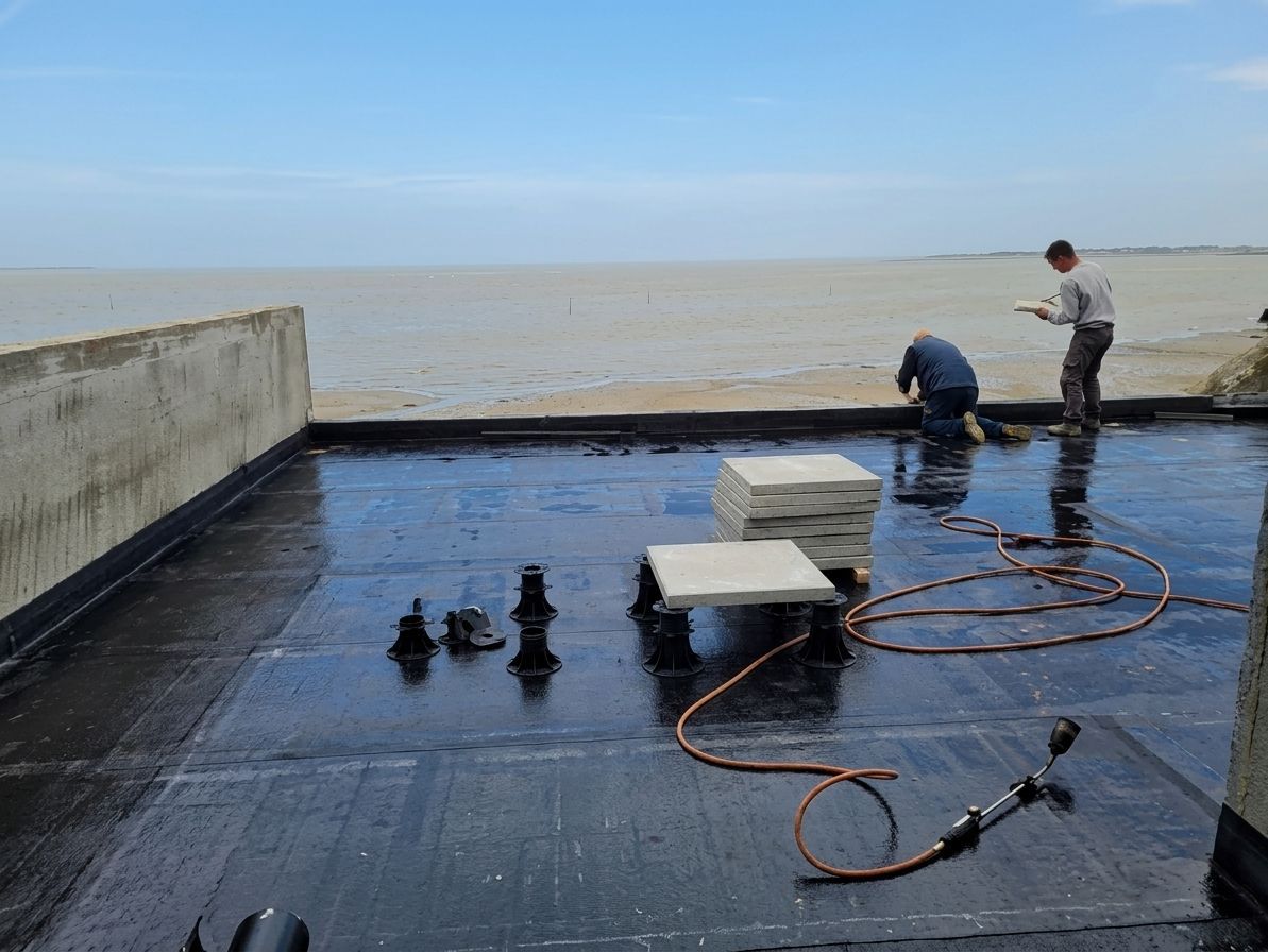 Des ouvriers réparent une surface noire étanche sur un toit plat près de l'océan.