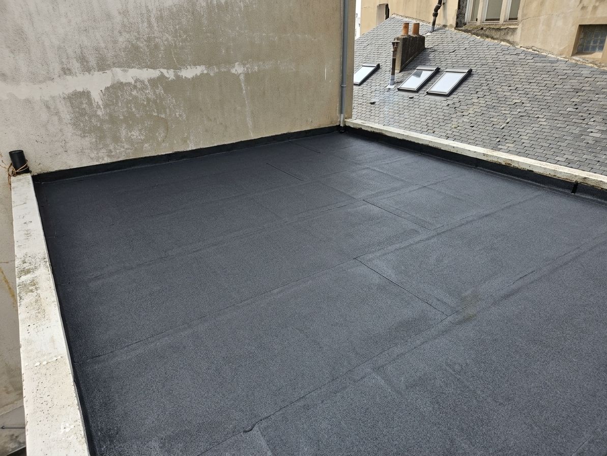 Toiture noire étanche jouxtant un mur beige, avec des bardeaux voisins visibles à proximité.