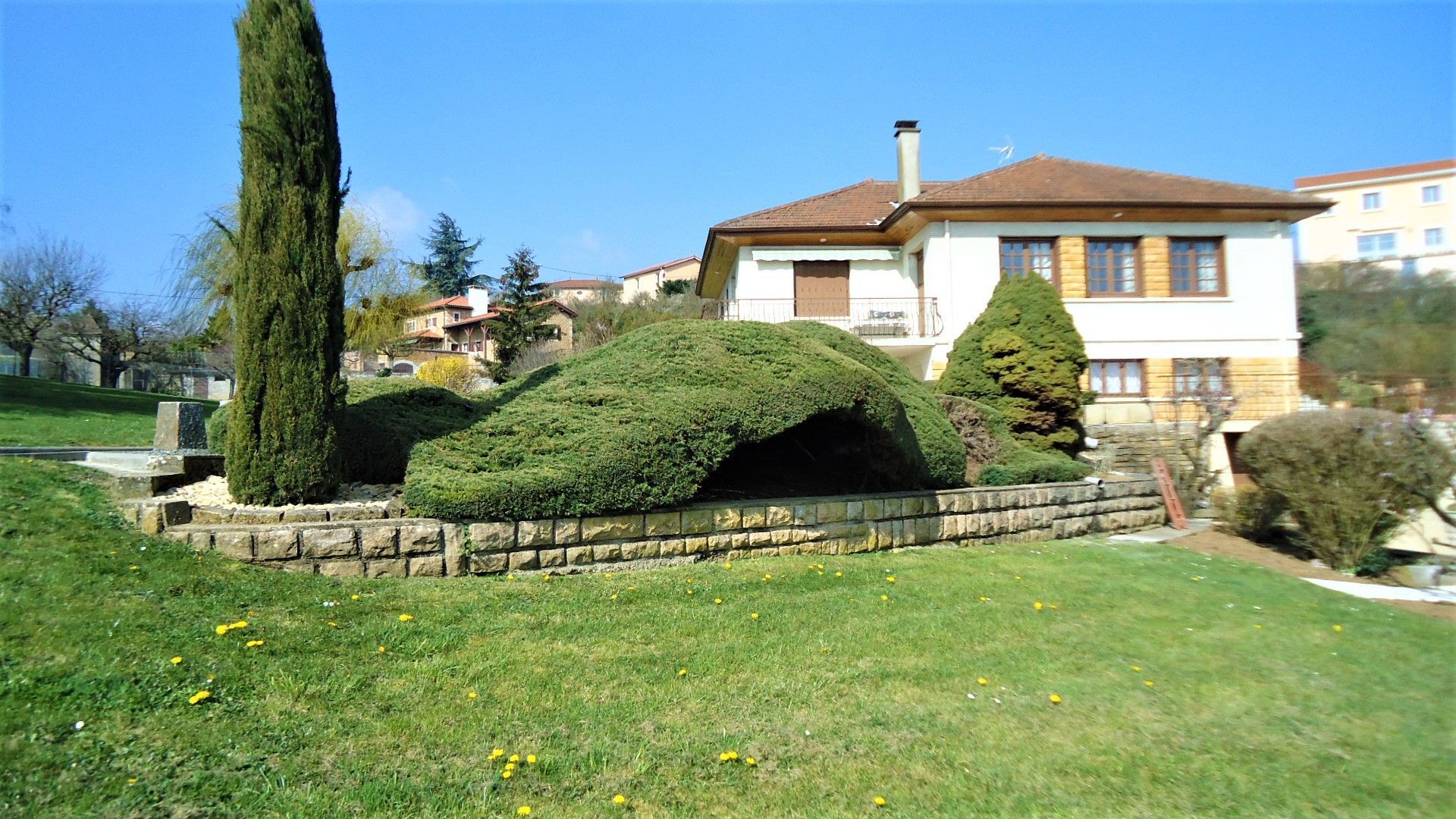 Une maison blanche au toit brun se dresse derrière un mur de soutènement en pierre et un jardin orné d'un grand arbuste à feuilles persistantes soigneusement taillé.