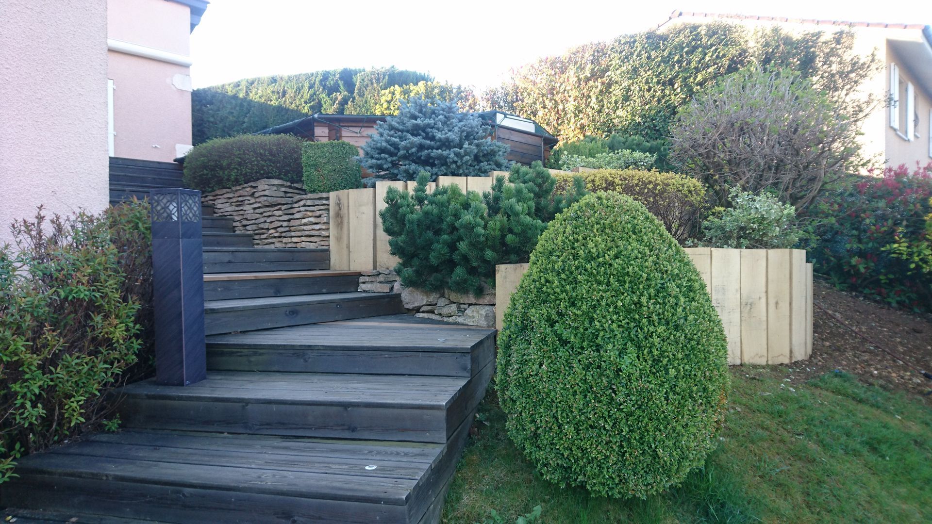 Un escalier extérieur en bois ; un buisson sphérique bien taillé ; des supports en bois clair.