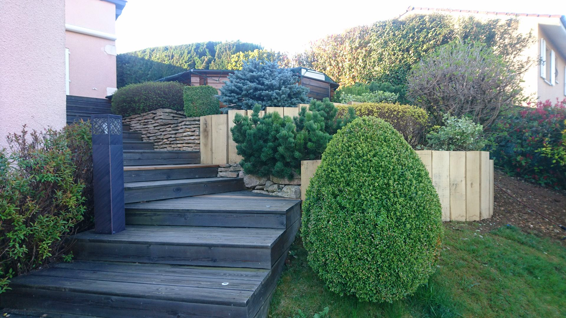 Des marches extérieures en bois sombre mènent à un chemin de jardin bordé d'arbustes taillés et de murs de soutènement en bois à plusieurs niveaux.