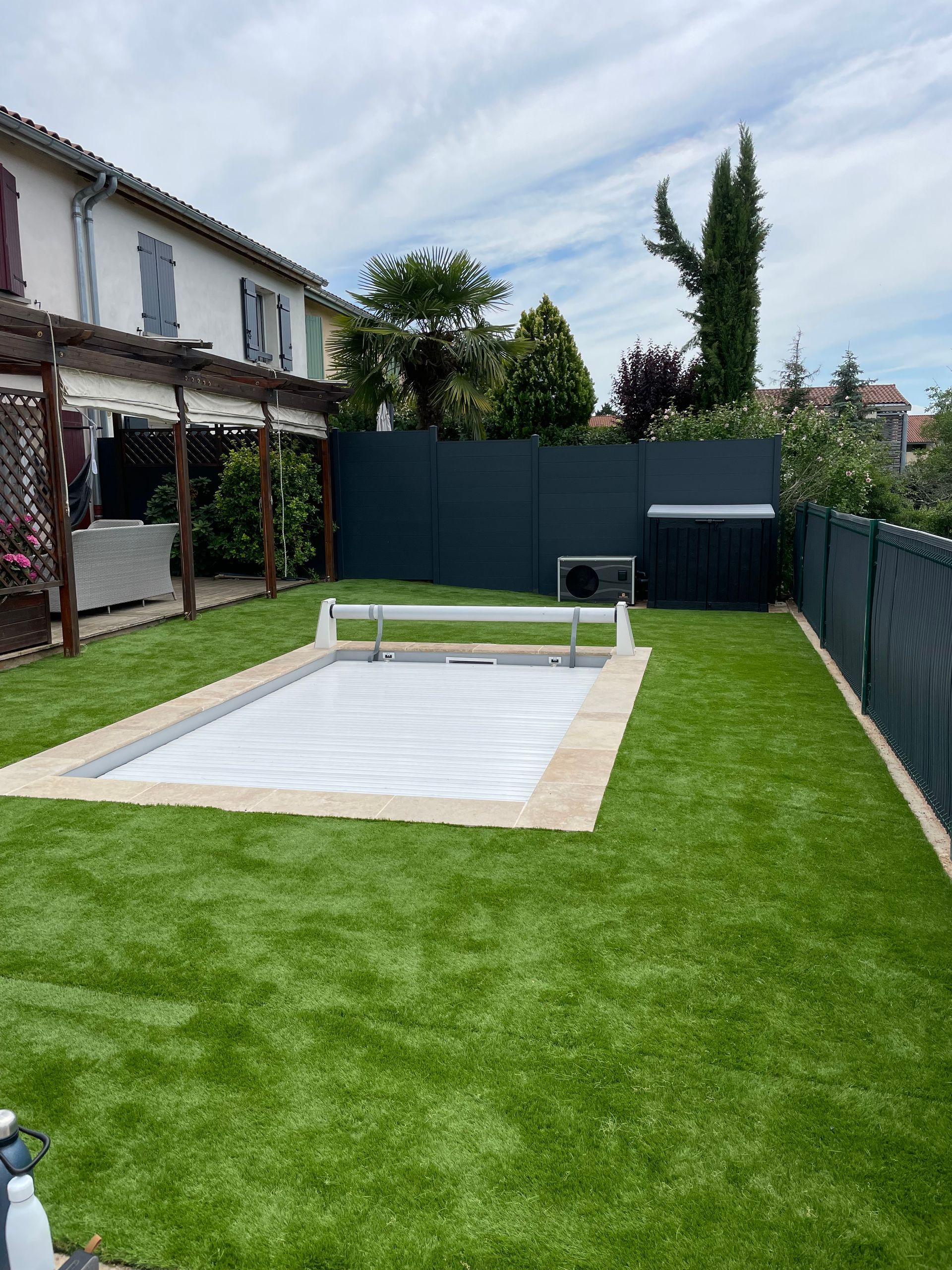 Un jardin avec une piscine rectangulaire recouverte d'une bâche solaire blanche, entourée de gazon artificiel et d'une clôture foncée.