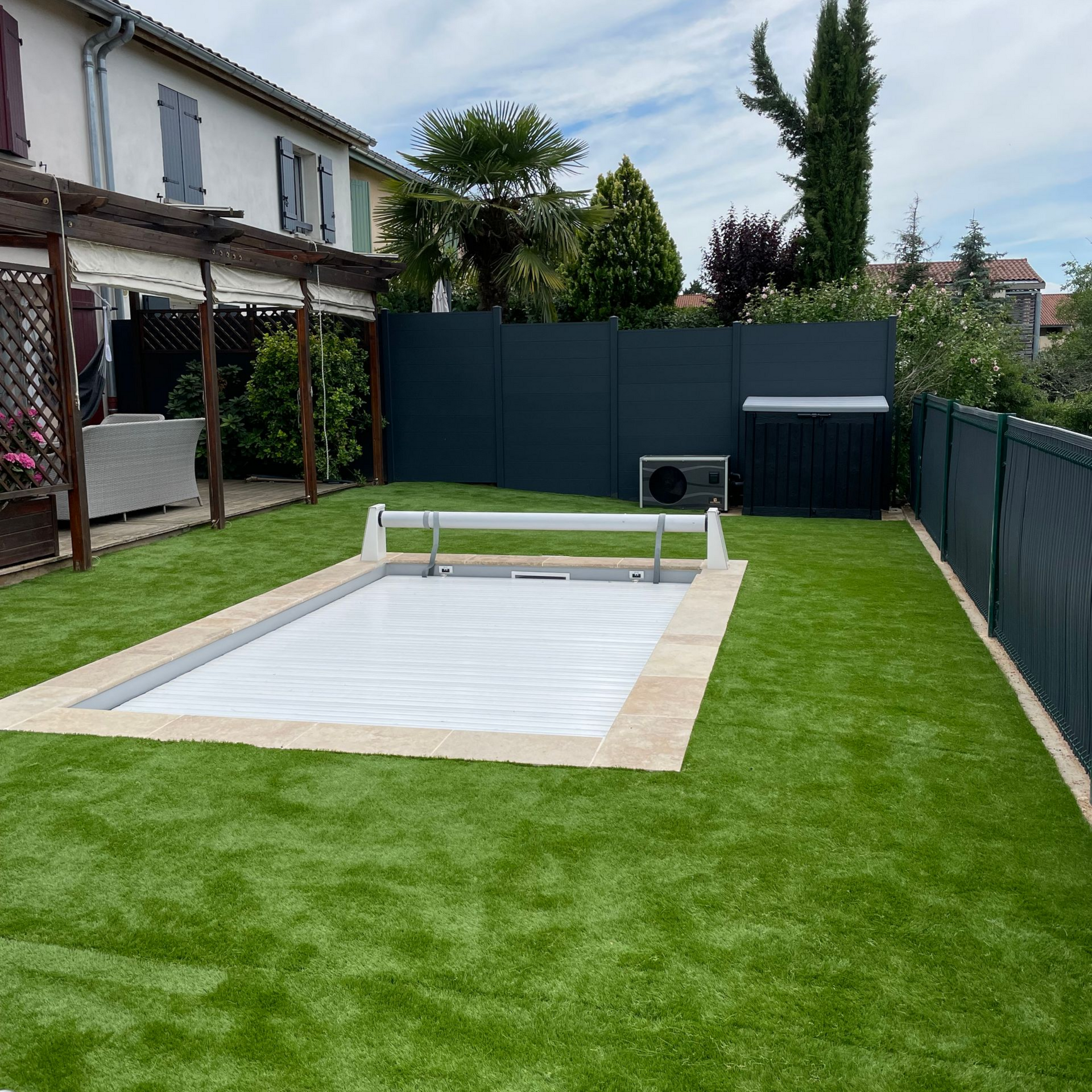 Une piscine de jardin avec une protection, entourée de gazon artificiel vert, d'une bordure en pierre beige et d'une clôture sombre pour préserver l'intimité.