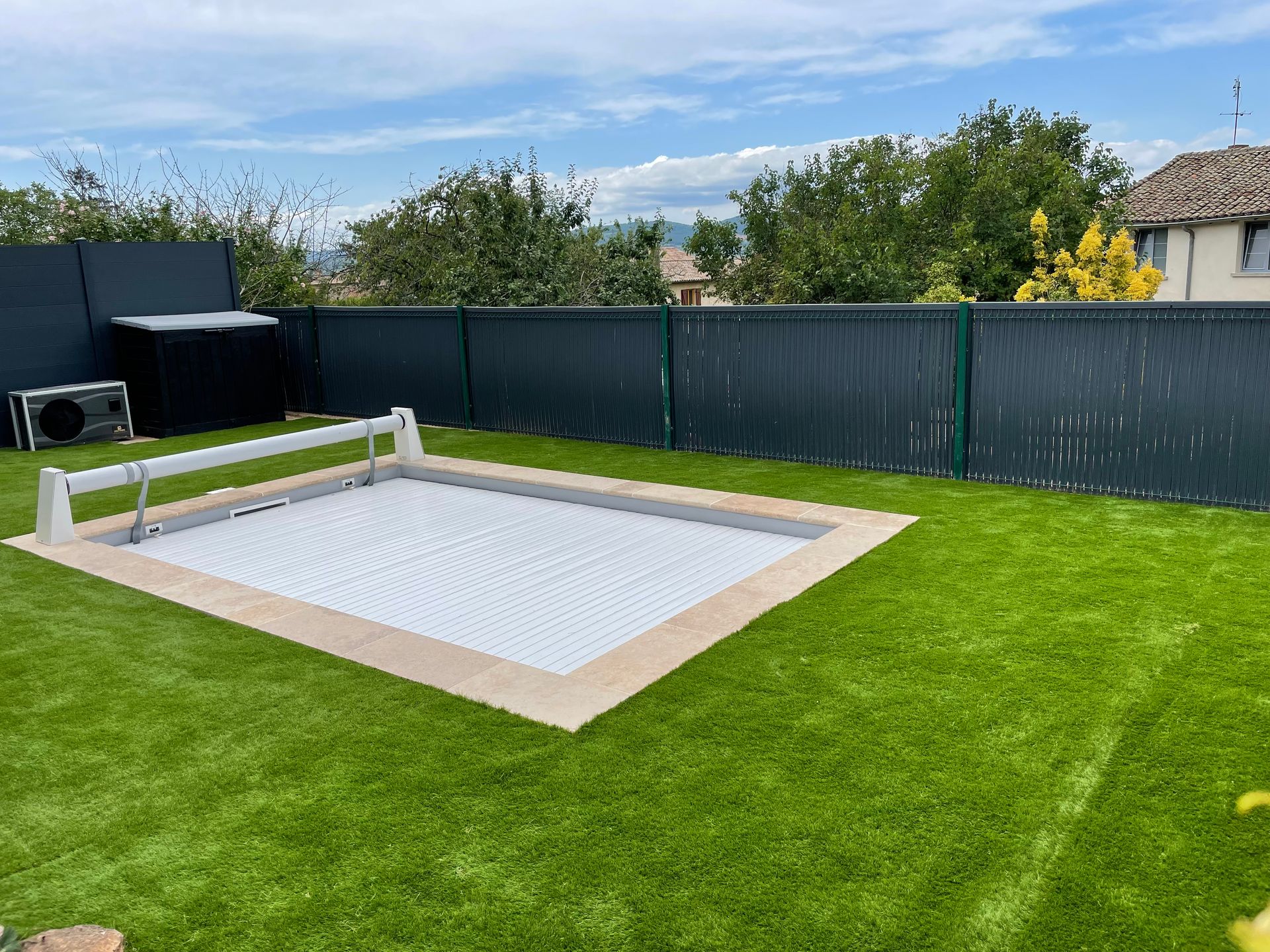 Une piscine rectangulaire dans un jardin, recouverte d'une bâche solaire gris clair, est aménagée sur une pelouse entourée d'une clôture préservant l'intimité.