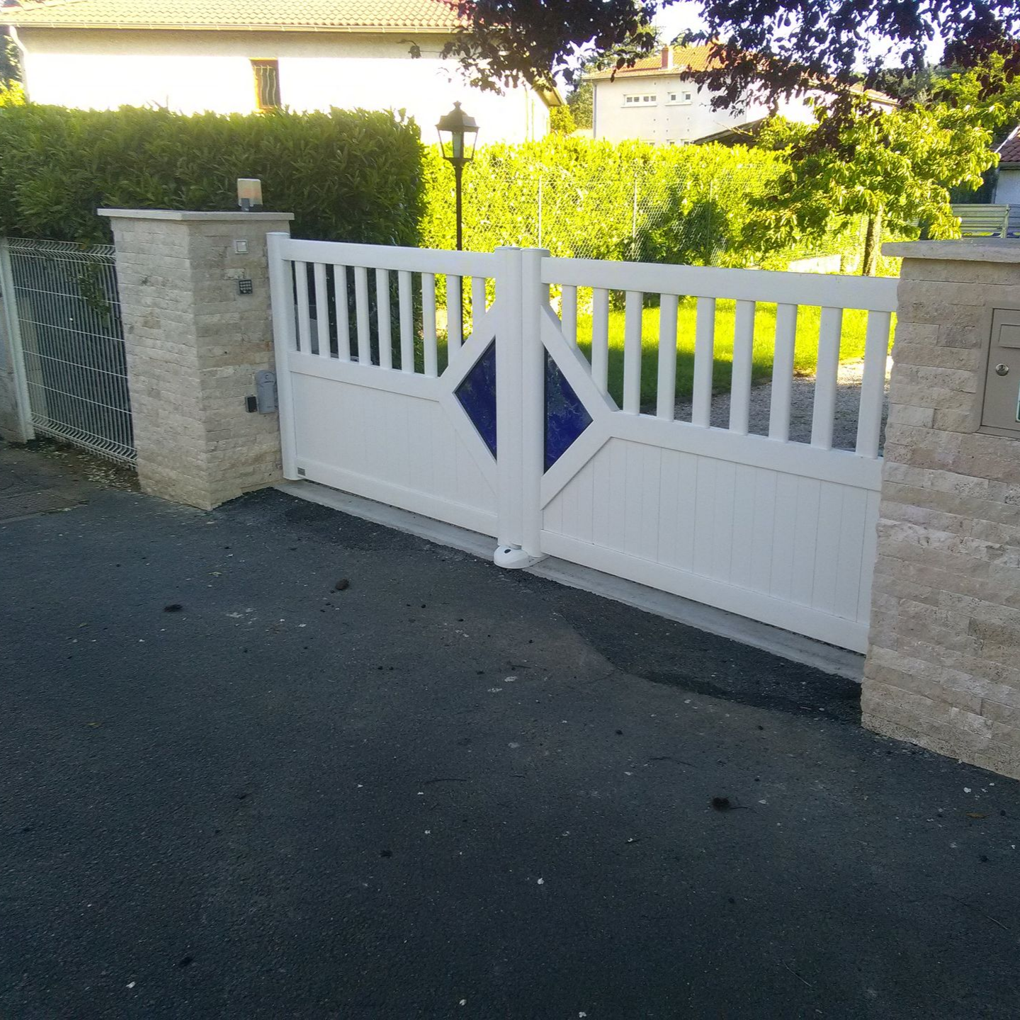 Un portail double blanc orné de motifs bleus en forme de losange, installé entre deux piliers de pierre devant une maison.