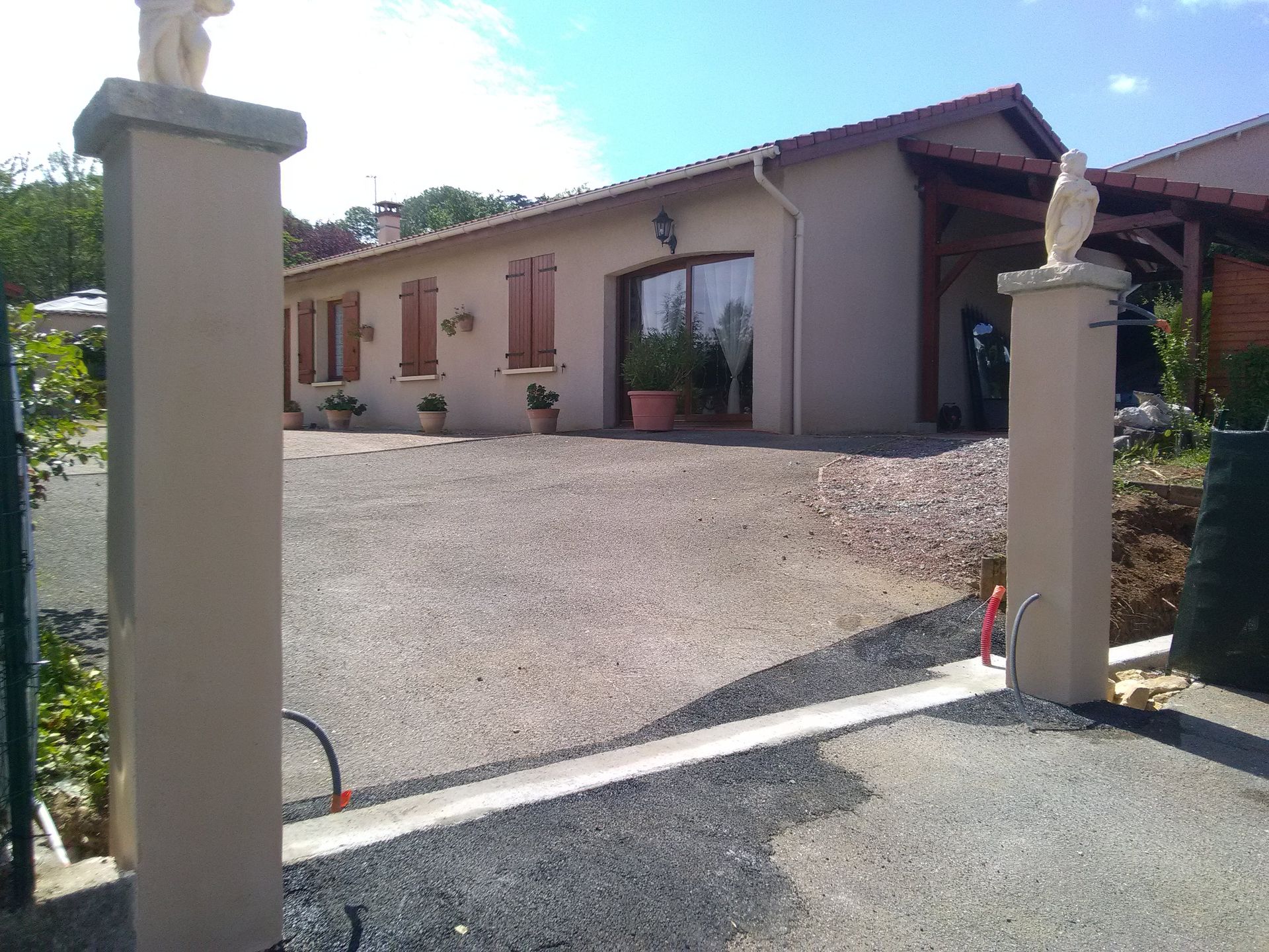 Une entrée encadrée par deux piliers de pierre surmontés de statues et une allée de gravier mènent à une maison de plain-pied de couleur claire.