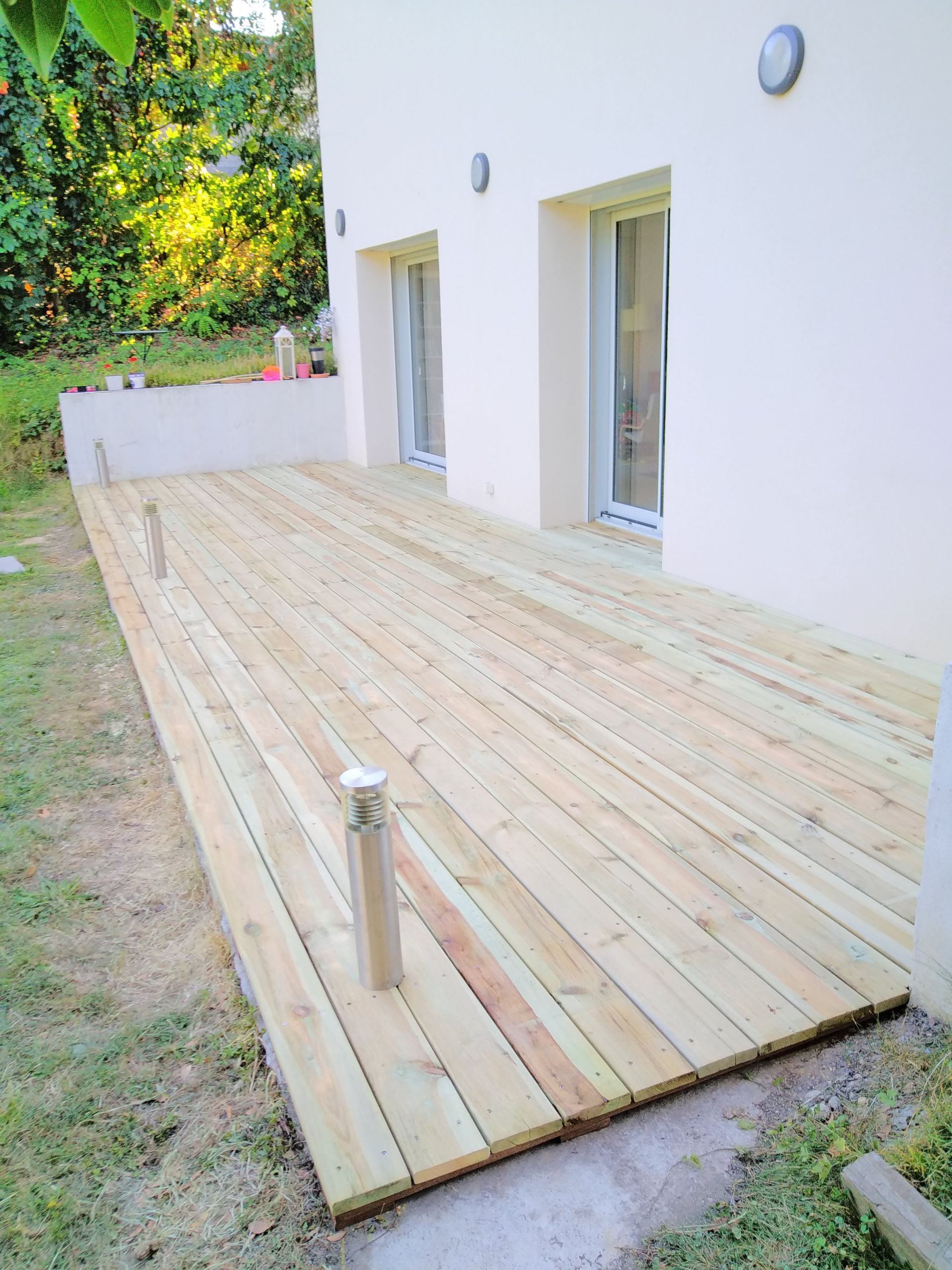 Une terrasse en bois installée le long d'un bâtiment de couleur claire, comportant deux portes extérieures et un éclairage au niveau du sol.
