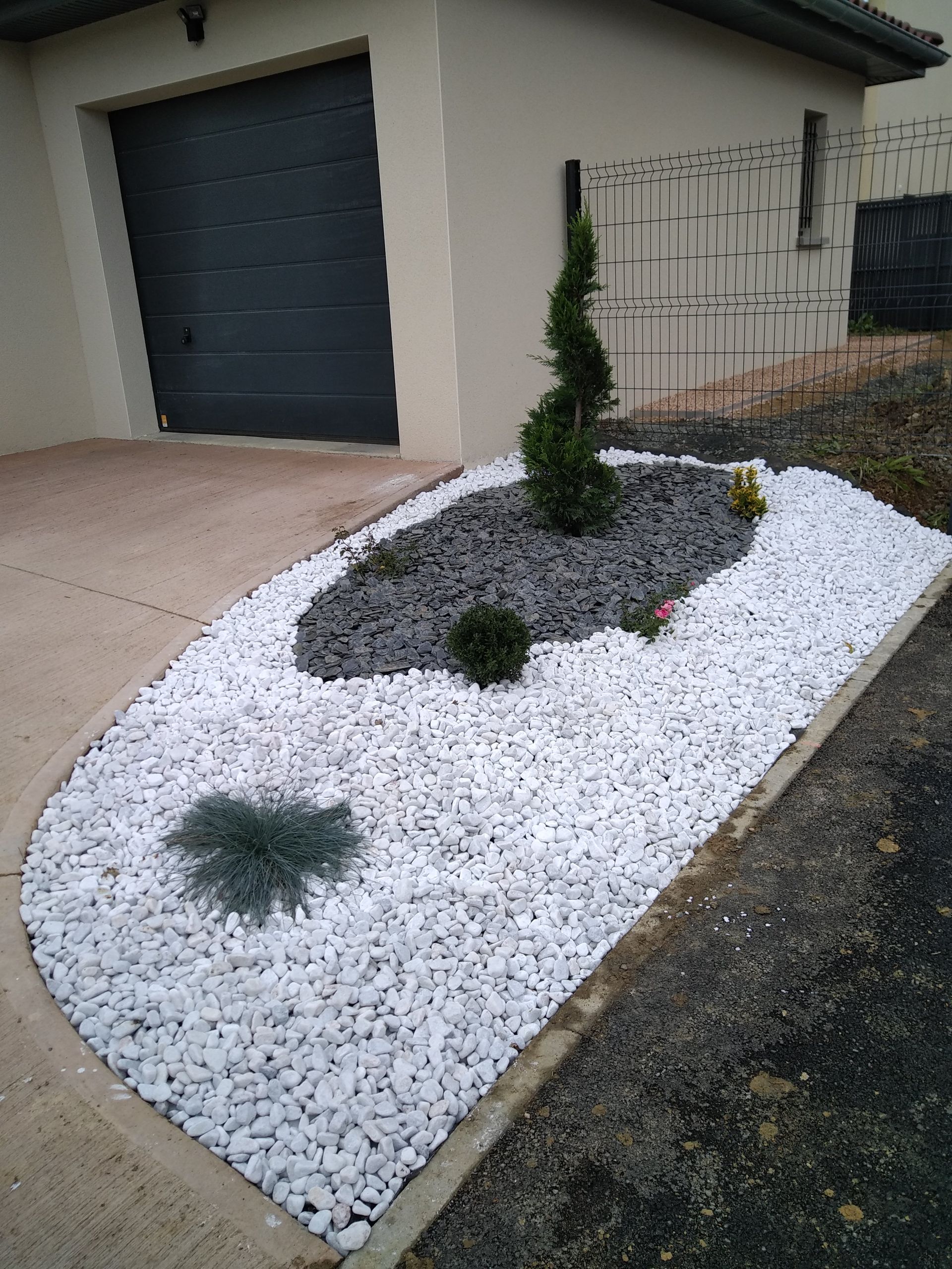 Un parterre de jardin aménagé avec un arbuste en spirale, un petit buisson et du paillis de pierre grise, entouré de gravier décoratif blanc.