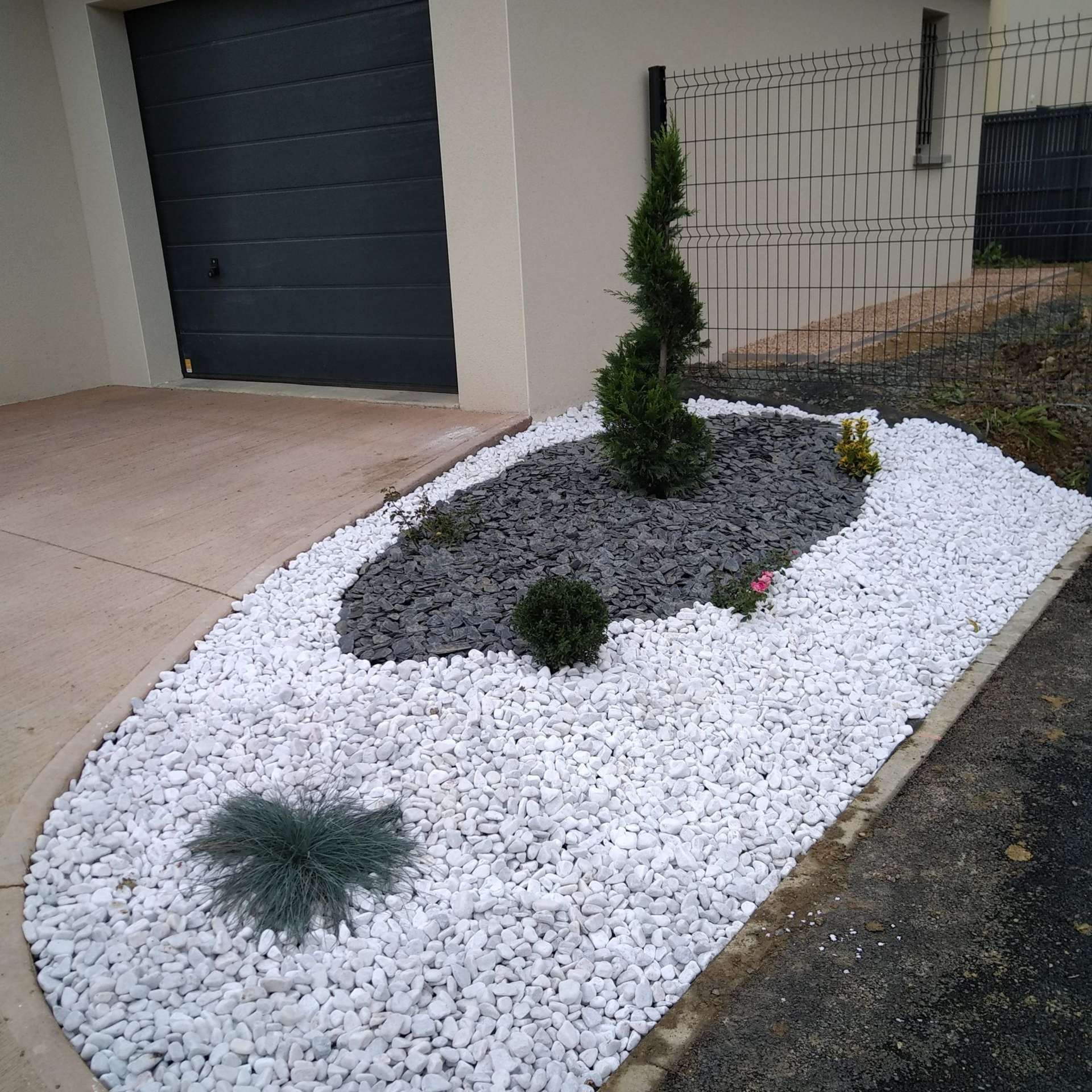 Un parterre de jardin aménagé avec de l'ardoise gris foncé au centre, entourée de galets blancs et un conifère taillé en spirale.