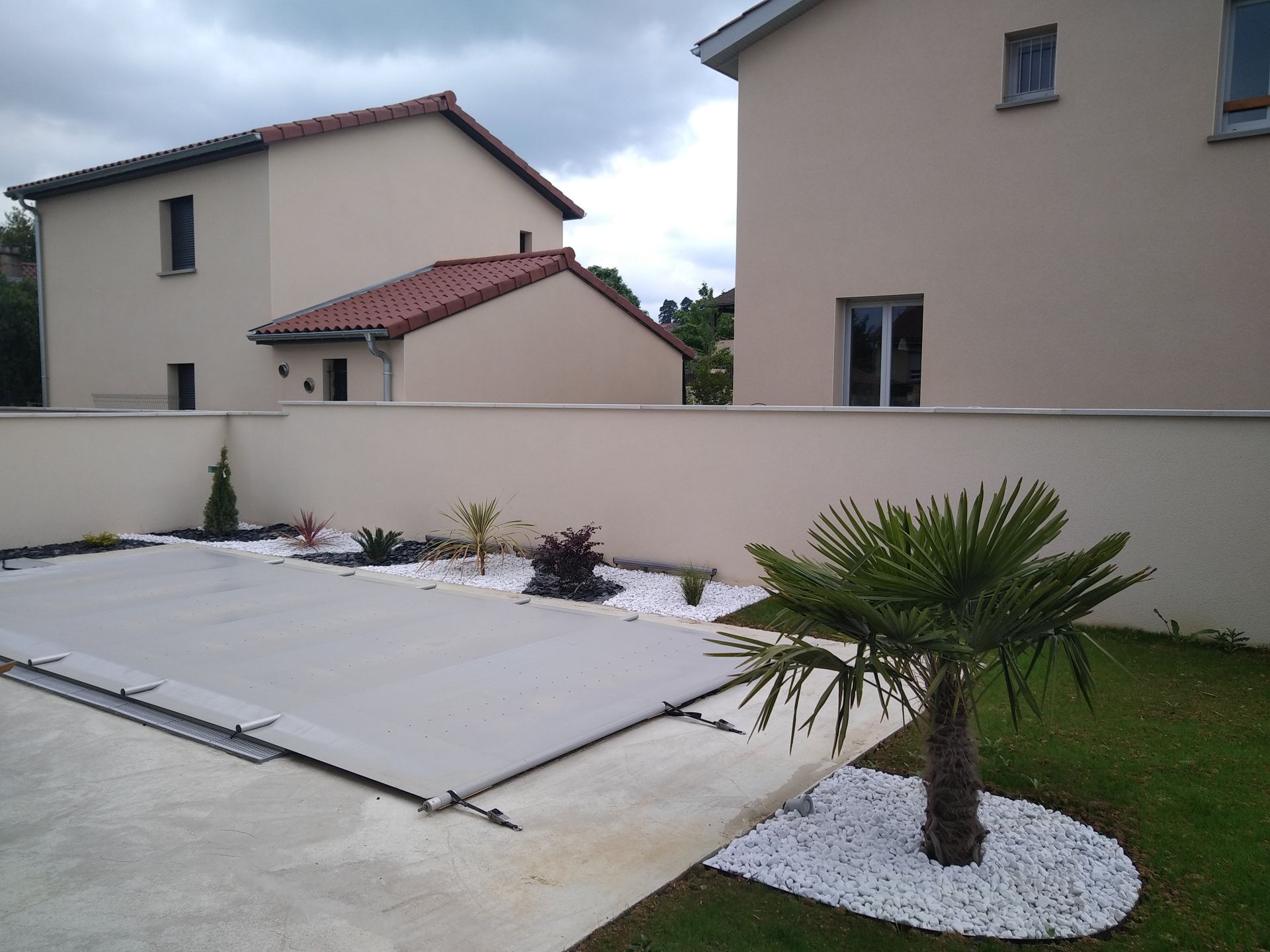 Une piscine recouverte d'une bâche grise côtoie un petit palmier et un parterre de gravier, le tout encadré par des maisons modernes.