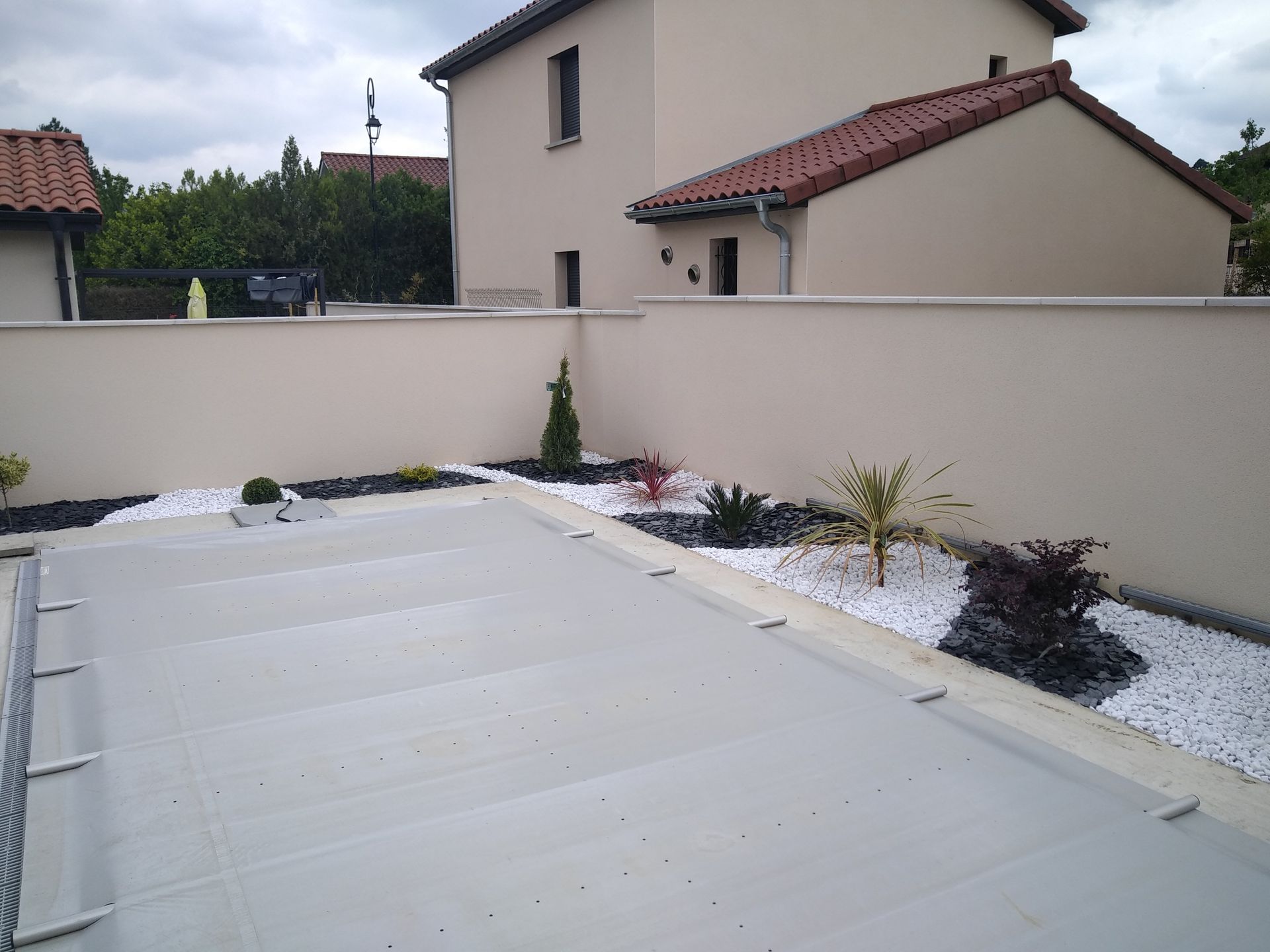 Une piscine recouverte d'une bâche de sécurité grise dans un jardin avec un mur beige, un aménagement paysager en gravier blanc et de petites plantes décoratives.