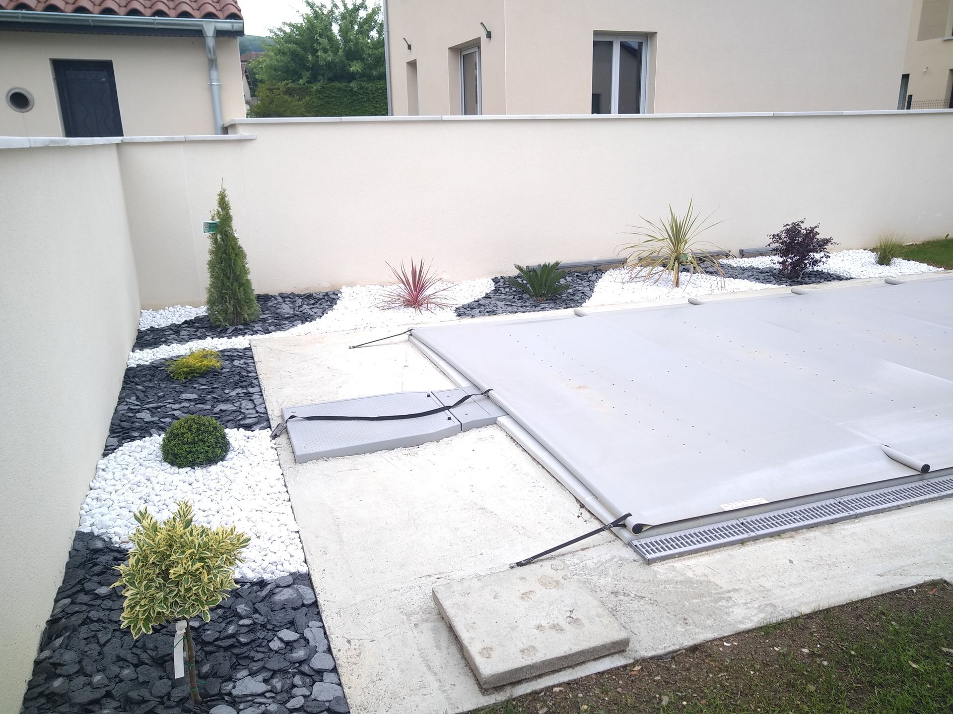 Une piscine de jardin recouverte d'une bâche grise, bordée de gravier décoratif blanc et gris foncé et de petits arbustes.