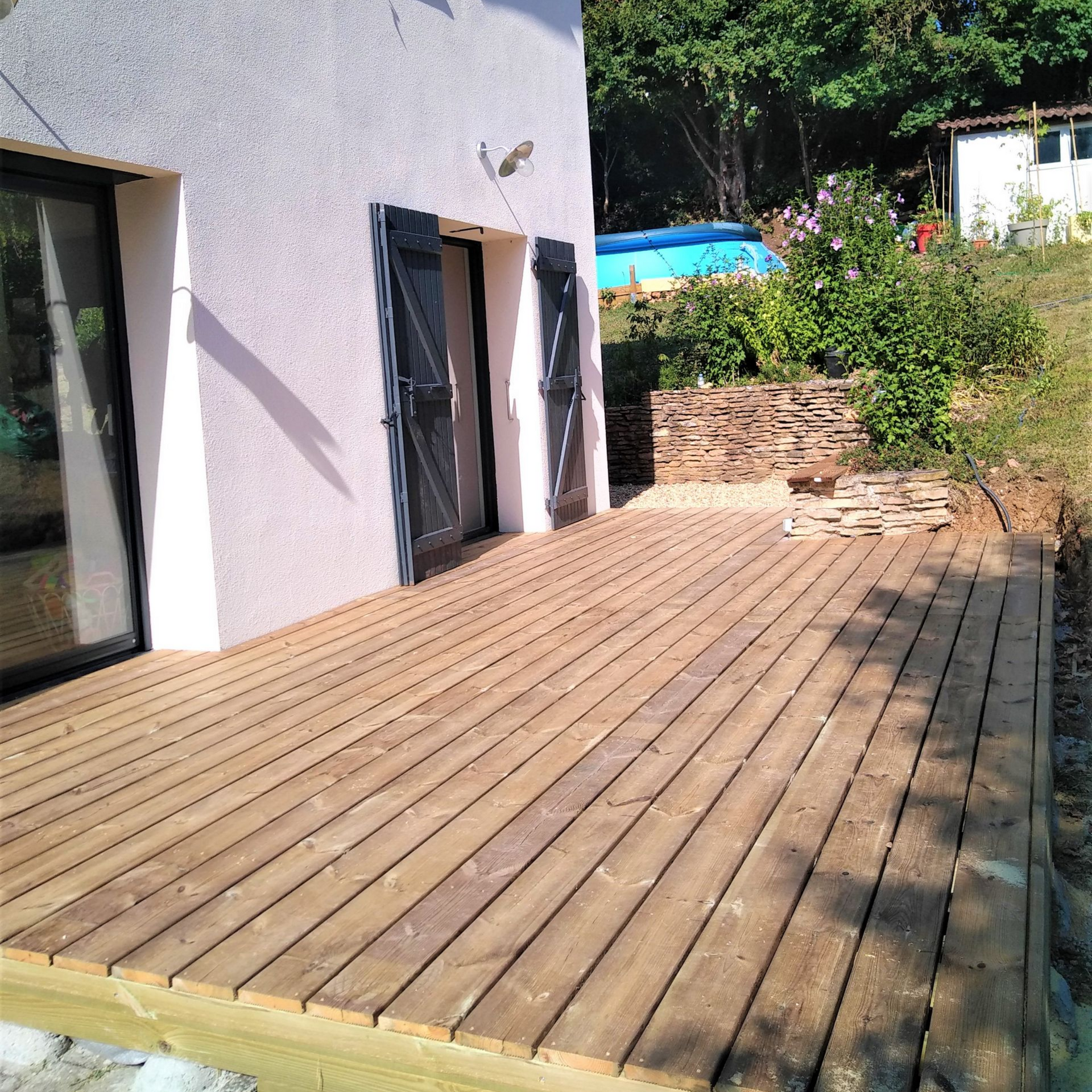 Une terrasse en bois attenante à une maison de couleur claire aux volets foncés, située à l'extérieur, à côté d'un jardin en pente et gazonné.