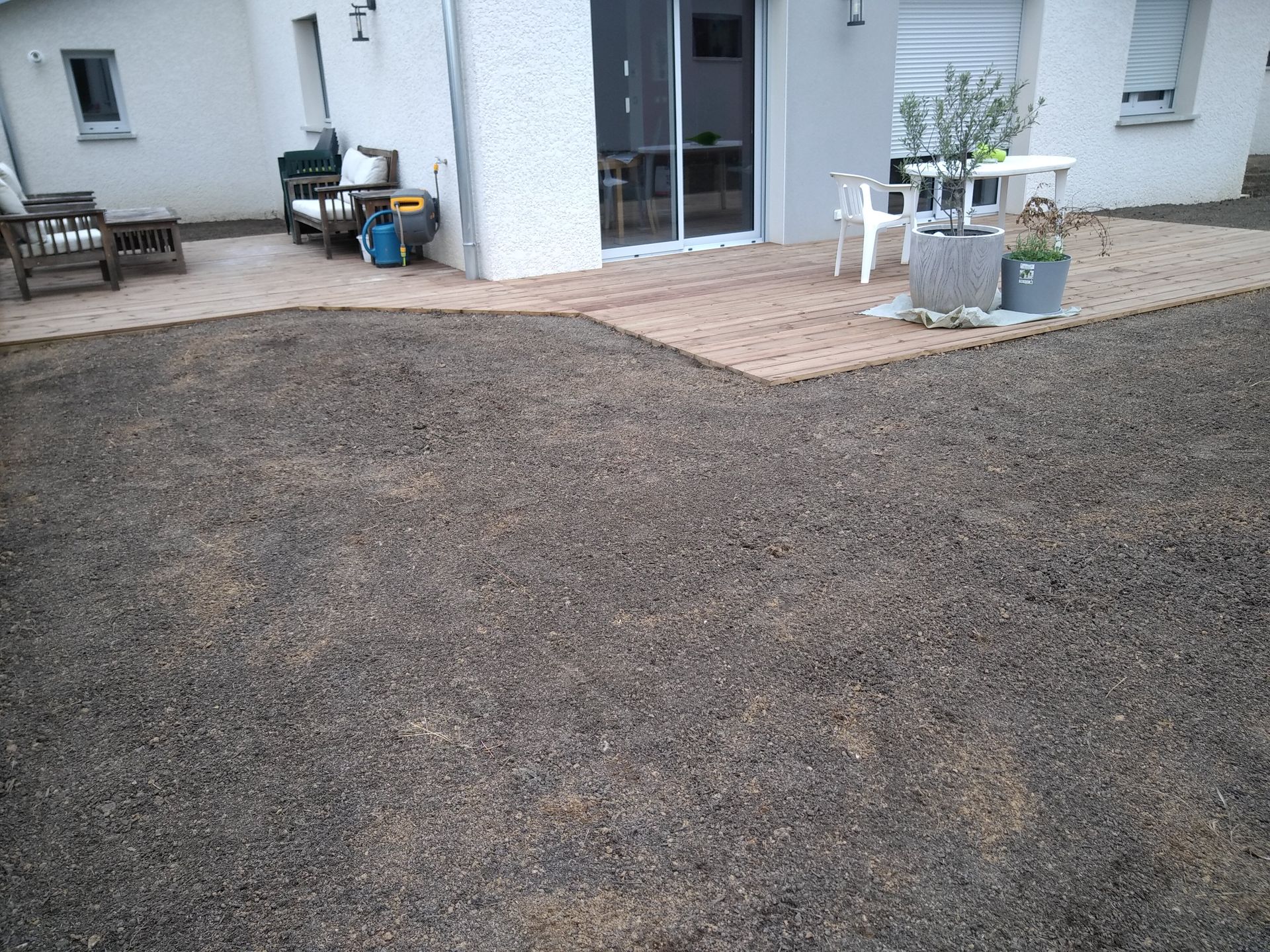 Une terrasse de maison en bois et du mobilier, bordant un grand espace en terre fraîchement nivelé.
