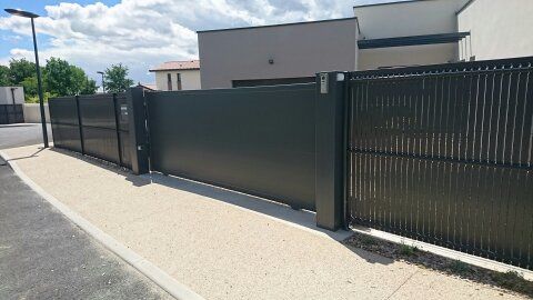 Un portail en métal gris foncé et une clôture assortie se dressent devant une allée de gravier clair et derrière une rue pavée.