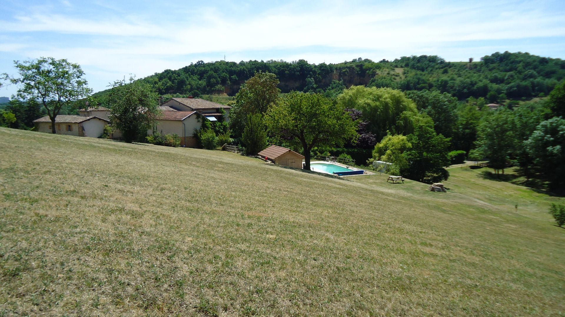Un versant herbeux descend en pente douce vers un groupe de bâtiments, une piscine et des arbres, avec une colline boisée à l'arrière-plan.