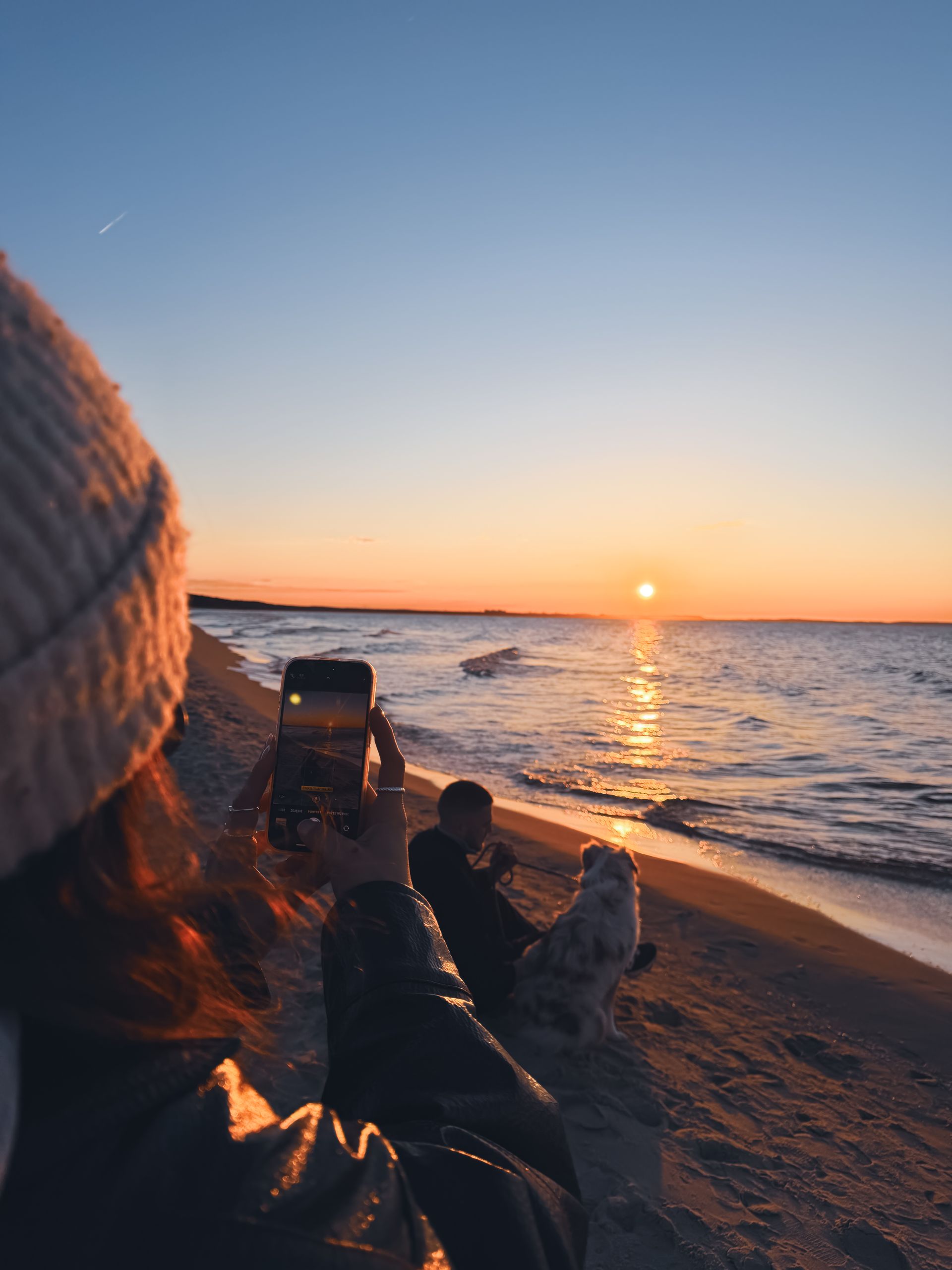 Osoba trzymająca telefon w celu sfotografowania zachodu słońca nad oceanem, z falami i piaszczystą plażą.