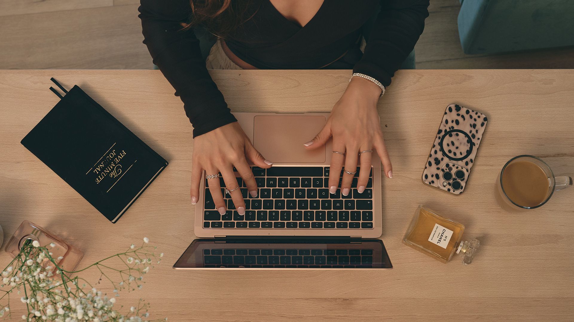 Dłonie piszące na laptopie przy drewnianym biurku z notatnikiem, telefonem, kawą i okularami.