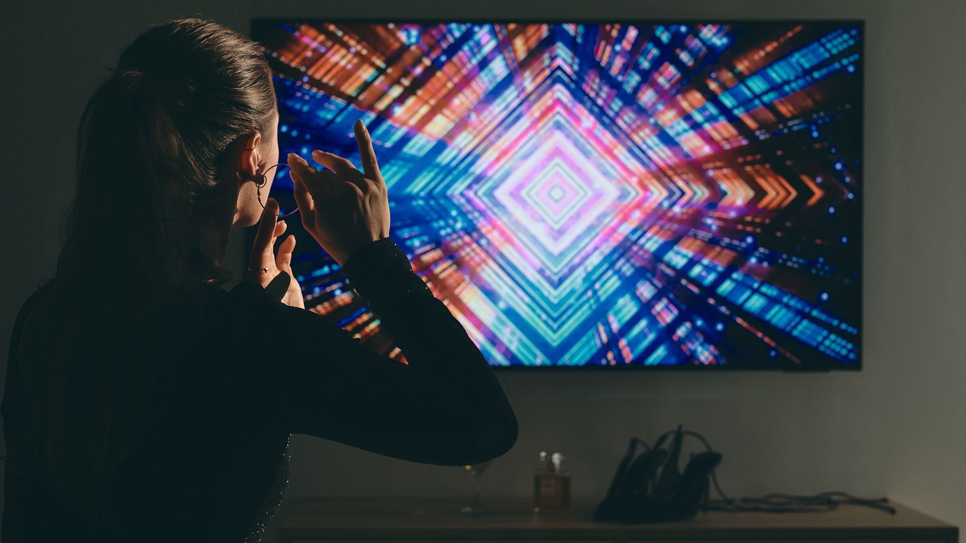 Osoba oglądająca telewizor zamontowany na ścianie z jasnym, neonowym wzorem geometrycznym w ciemnym pokoju