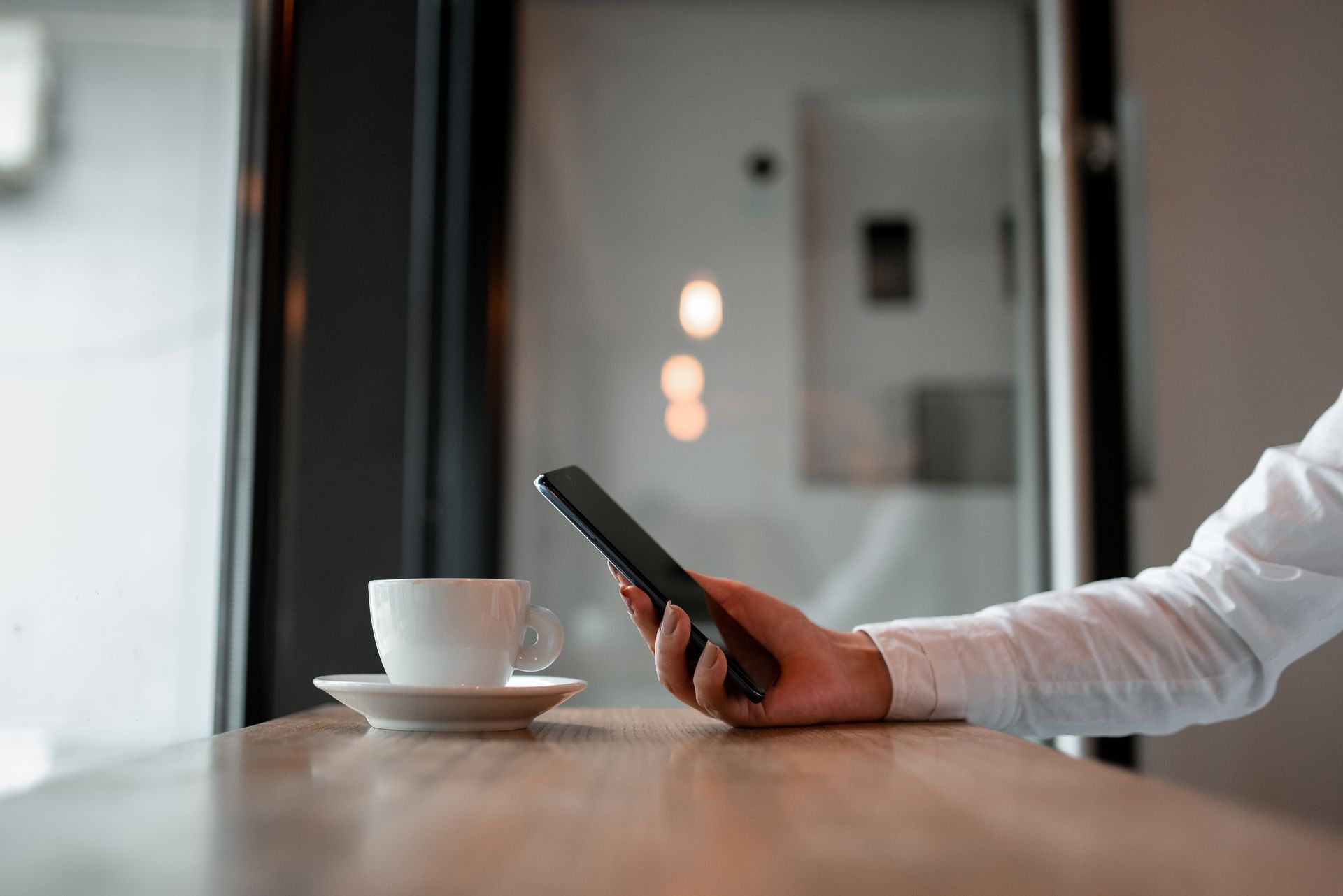 Ręka trzymająca smartfon obok białego kubka na drewnianym stole w nowoczesnym pokoju