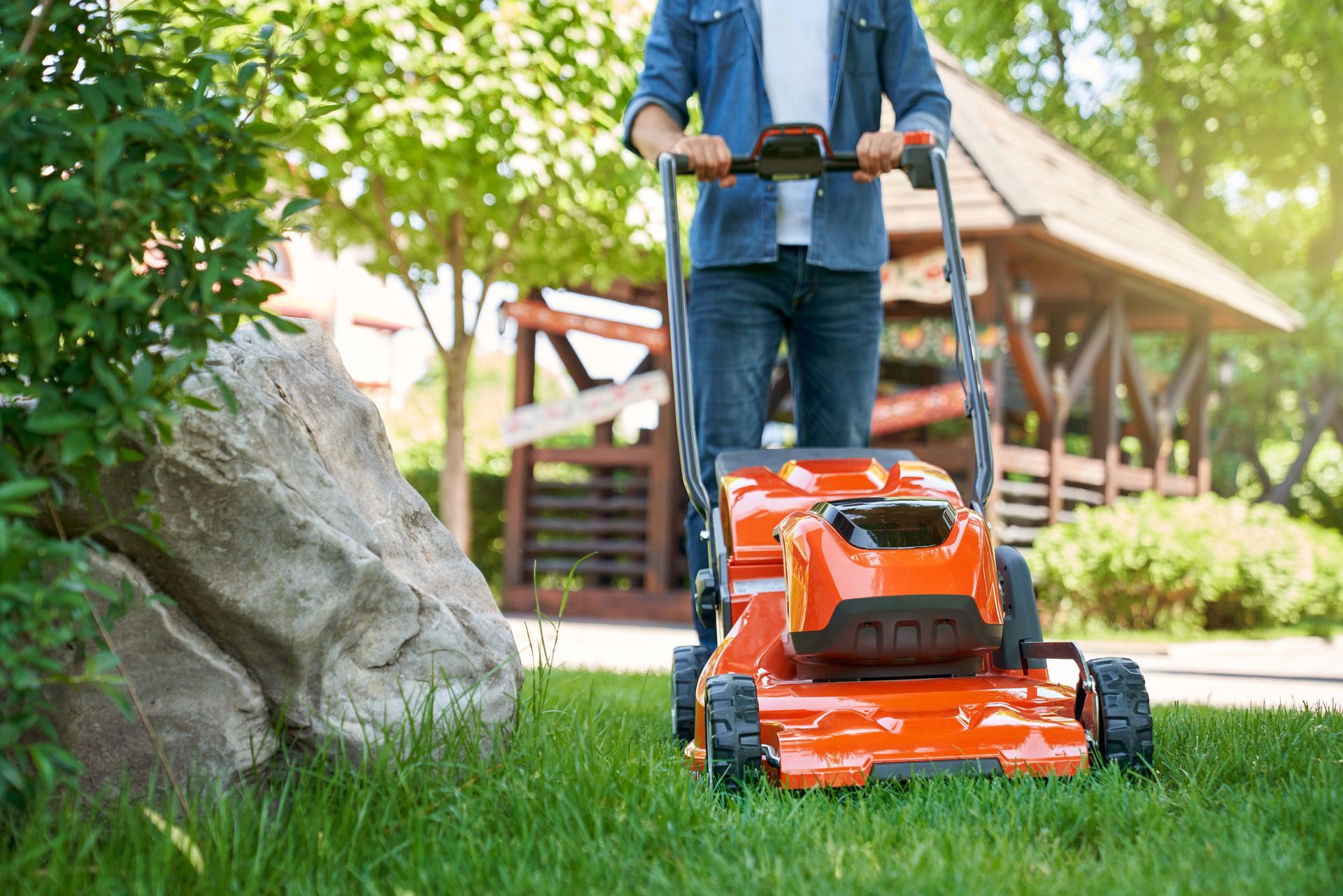 Une personne tond une pelouse avec une tondeuse électrique orange dans une cour ensoleillée avec un rocher et un belvédère.