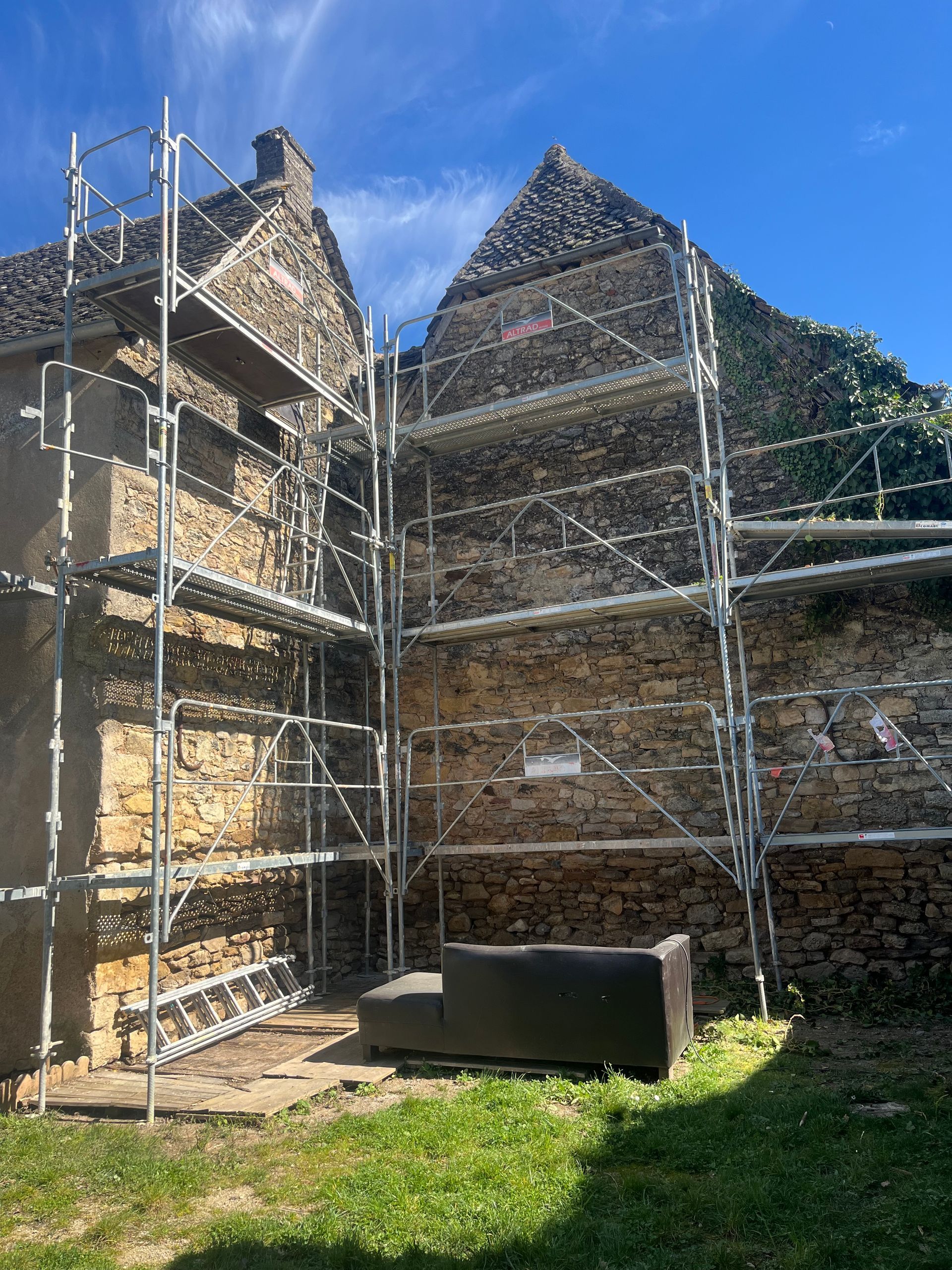 Échafaudage contre un bâtiment en pierre en cours de rénovation ; un canapé est posé en dessous. Ciel bleu éclatant.